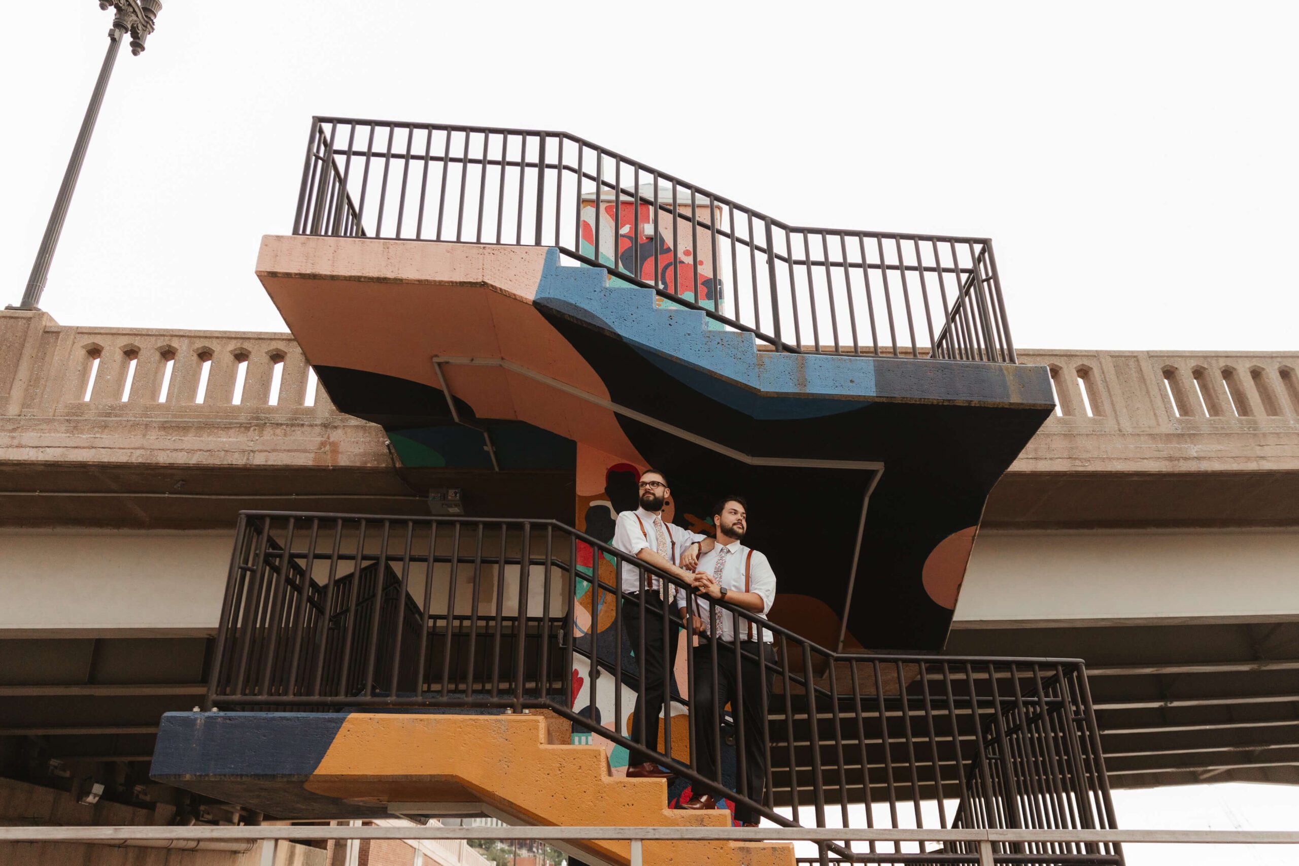 Two people stand on a colorful, geometrically painted staircase outdoors, gazing into the distance. The stairway features bold patterns in orange, black, blue, and beige, and is surrounded by metal railings.