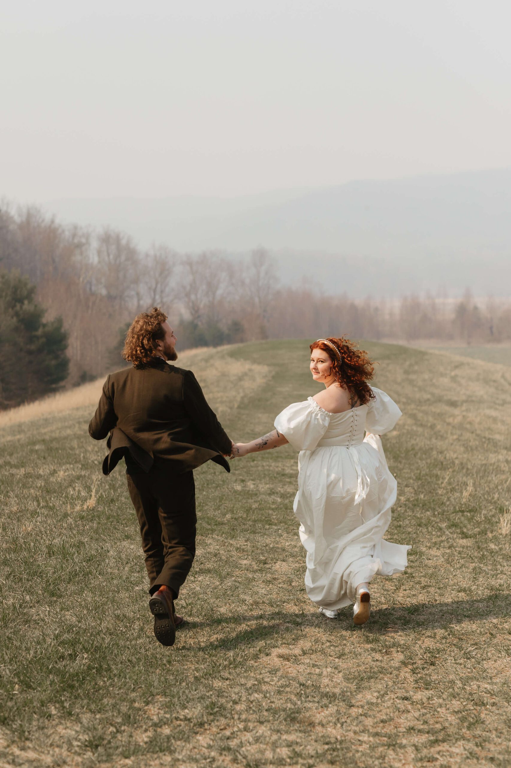 A couple in formal attire, with the woman in a white dress and the man in a dark suit, joyfully run hand in hand across a grassy field. The background features soft-focus trees and a hazy sky, suggesting a serene and romantic setting.