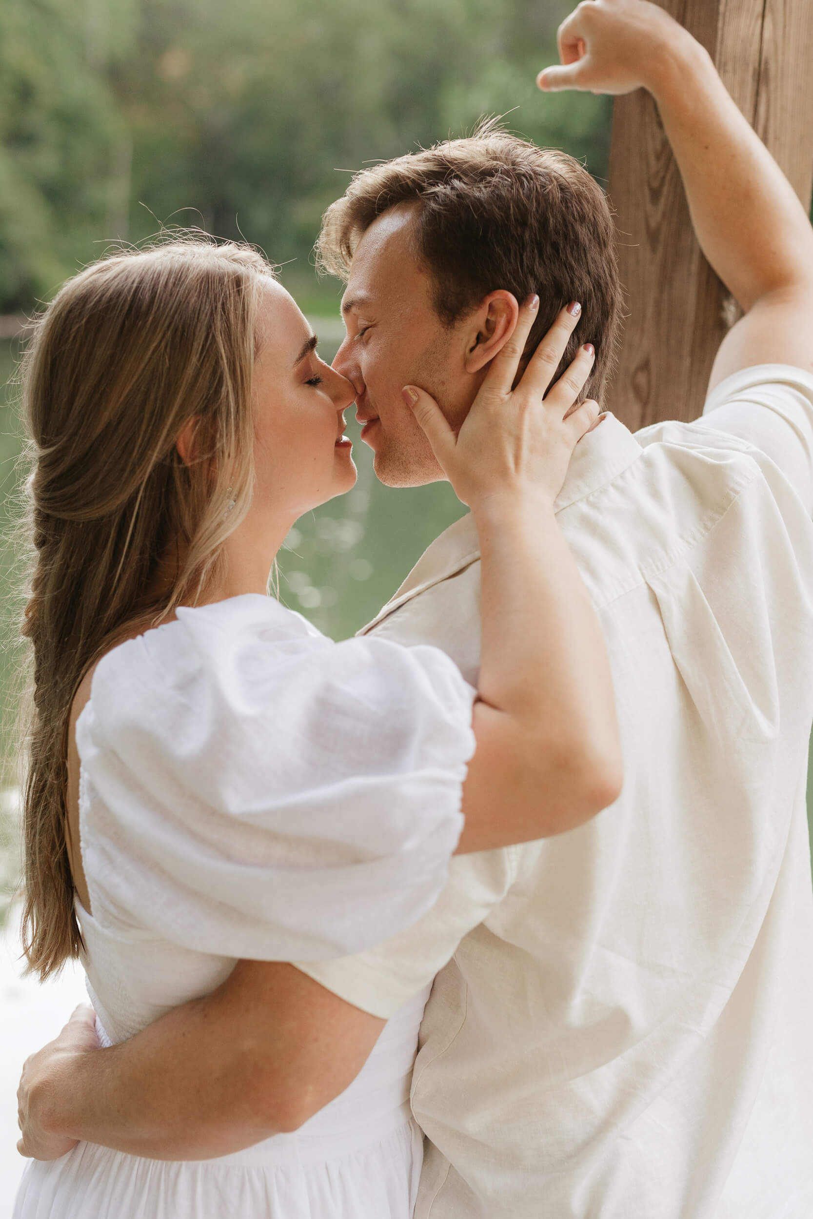 A couple stands close together beside a wooden post in an outdoor setting. They are gazing into each others eyes and about to kiss. Both are wearing light-colored clothing, and greenery is visible in the background.