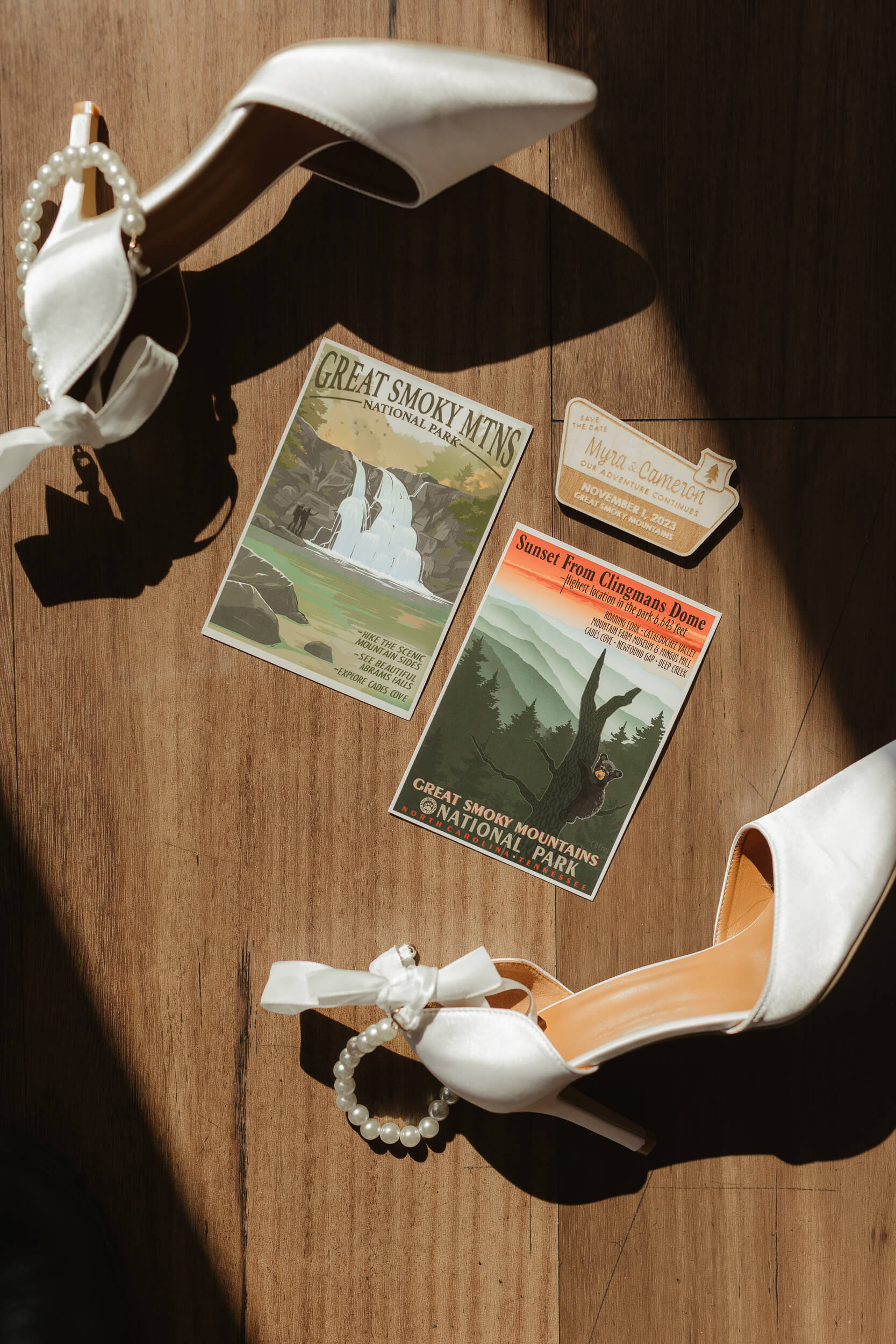 White high-heeled shoes with pearl anklets are placed on a wooden floor. Next to them are vintage-style postcards featuring the Great Smoky Mountains National Park, along with a small cutout shaped like a luggage tag. Pricing by Erin Morrison Photography.