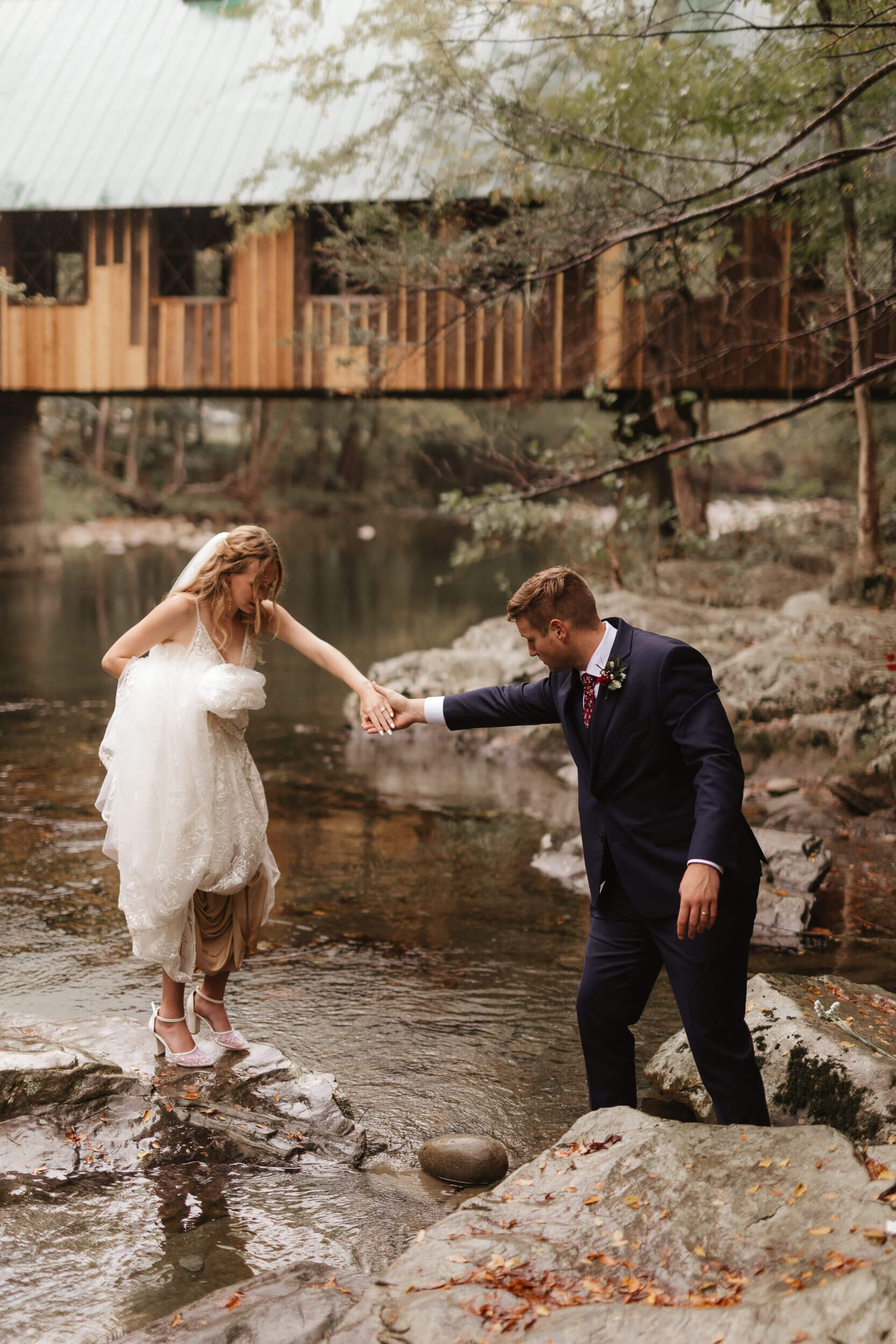 A bride and groom navigate stepping stones in a creek. The groom holds the brides hand, assisting her across. A wooden bridge and lush trees are visible in the background, creating a rustic outdoor setting.