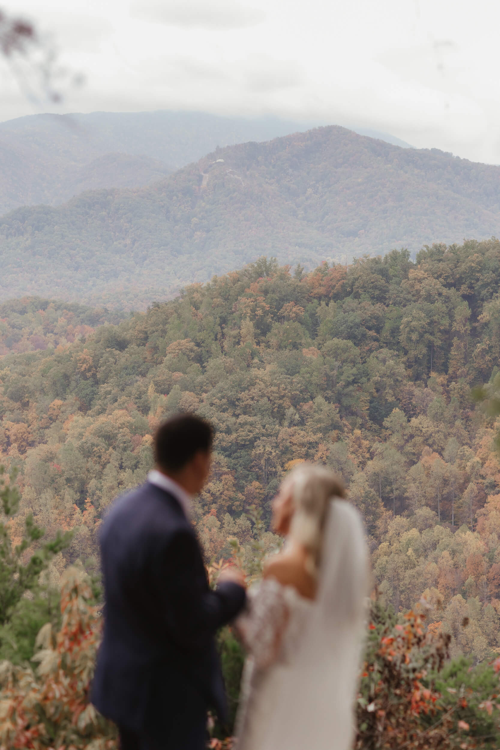 A couple in wedding attire stands in front of a scenic mountain view. The trees display autumn colors, creating a picturesque backdrop. The couple is slightly out of focus, emphasizing the natural beauty surrounding them.