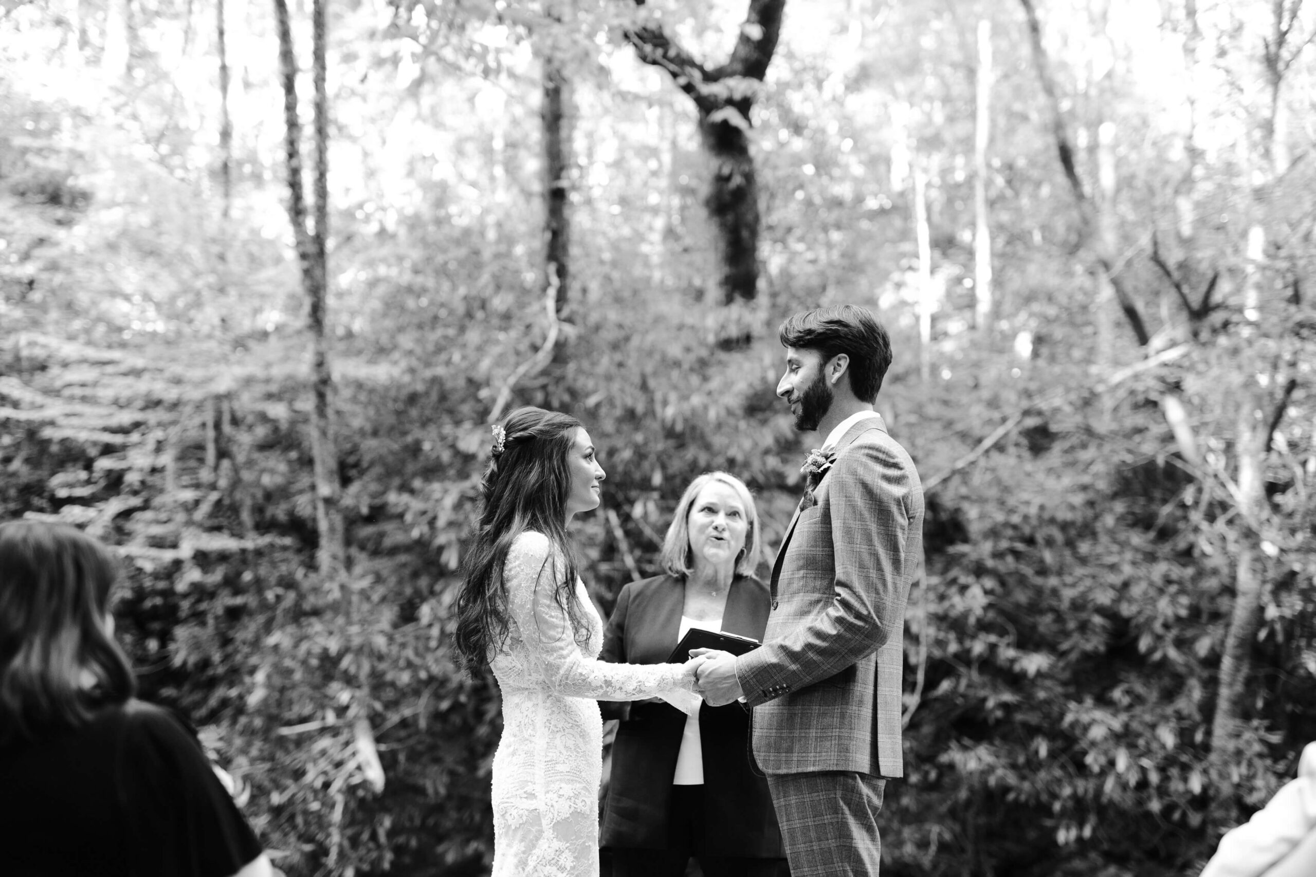 A black and white photo of a couple holding hands during a wedding ceremony in a forest setting. The bride is in a lace dress, and the groom is in a suit. A person officiates the ceremony under a tree canopy.