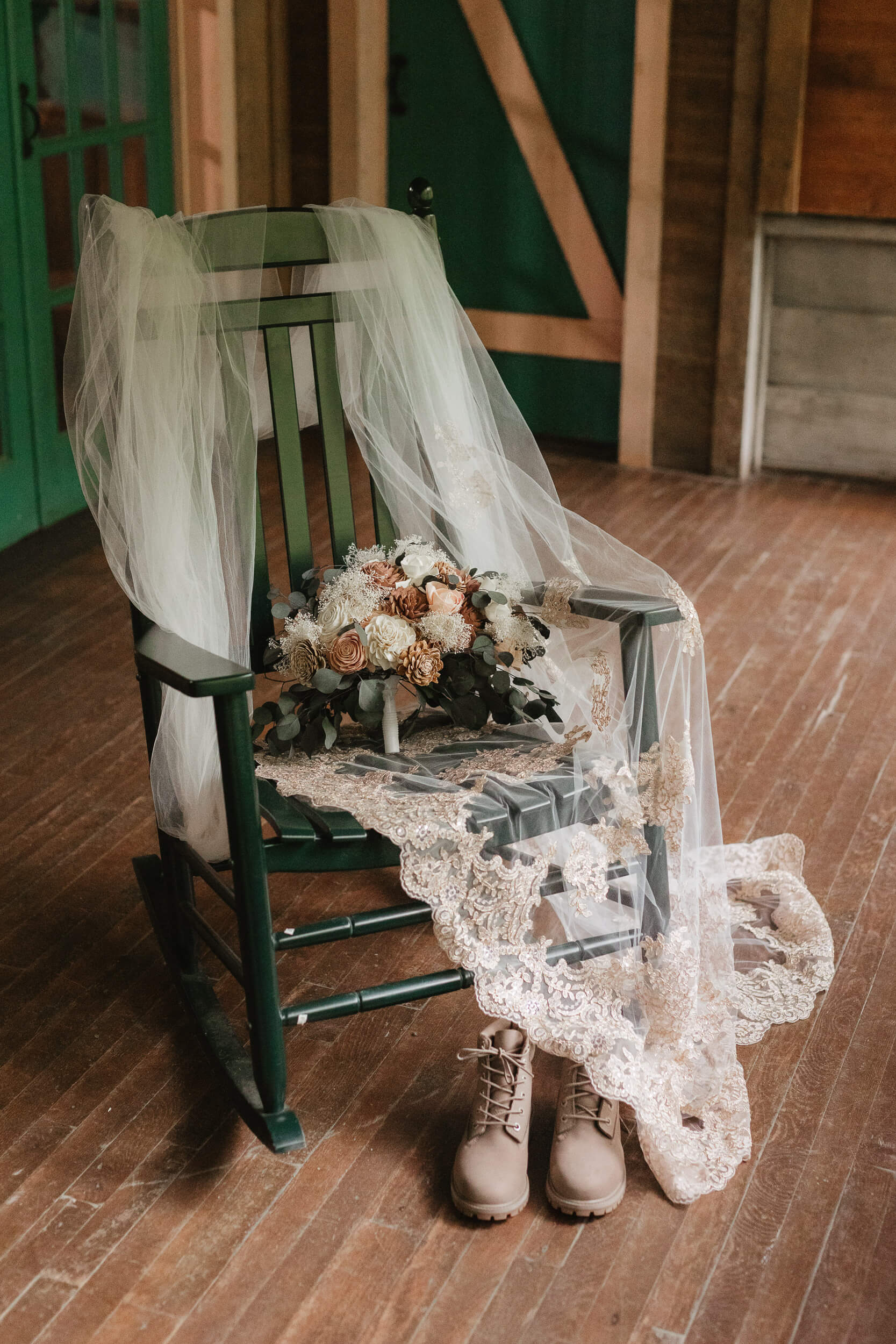 A dark green rocking chair draped with a delicate white lace veil. A bouquet of flowers rests on the seat, and a pair of tan boots is placed on the wooden floor nearby. The setting appears rustic and cozy.