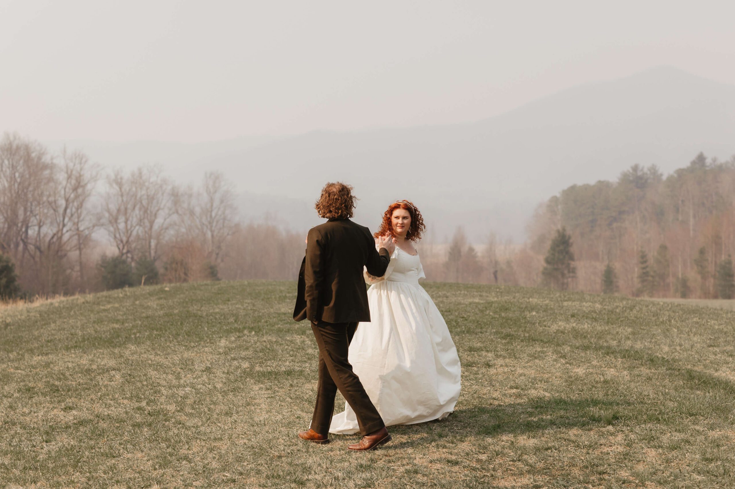 A couple dances together outside on a grassy field. The woman wears a white wedding dress, and the man wears a dark suit. Bare trees and distant mountains create a misty backdrop.