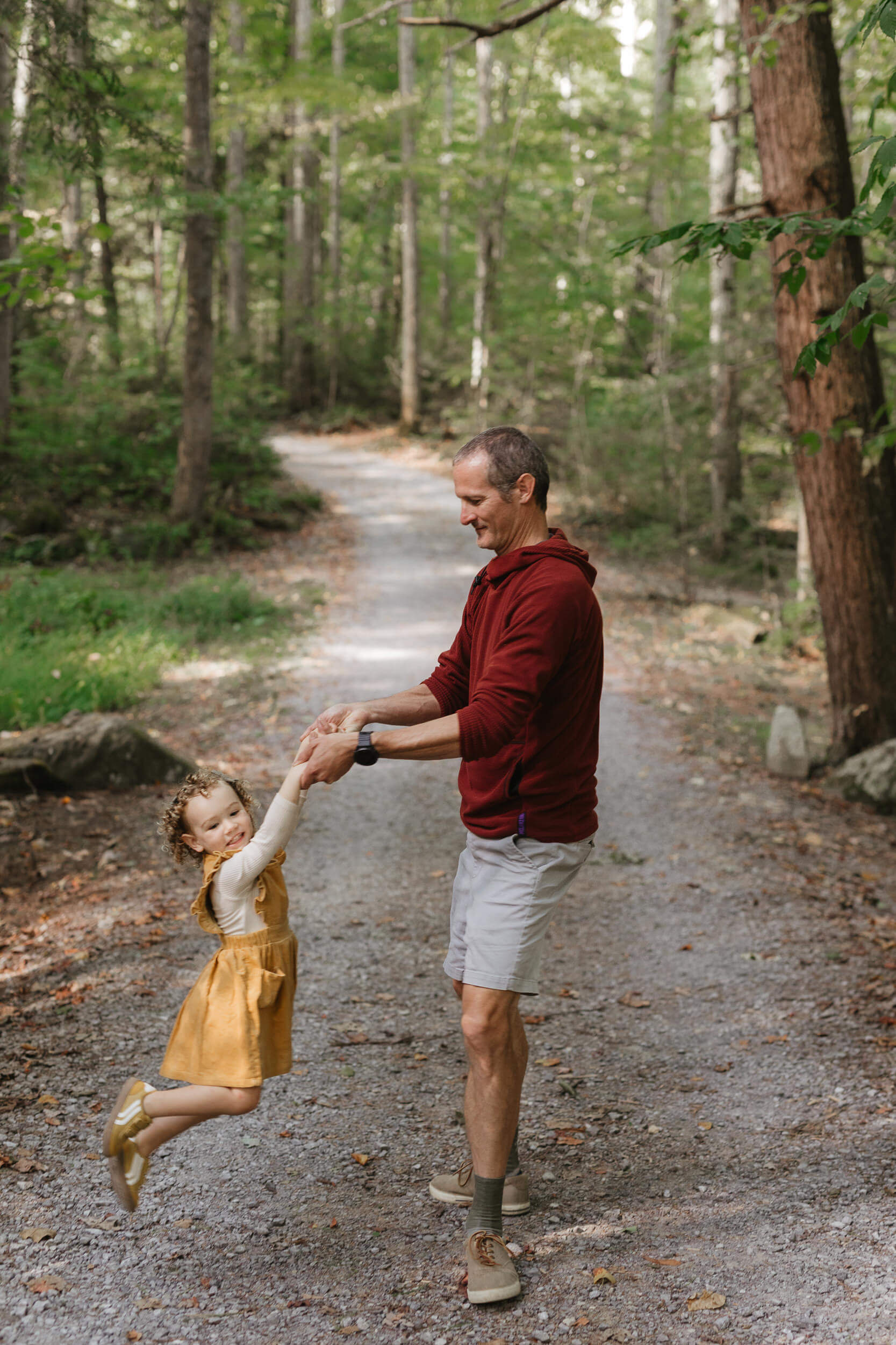 A man in a red hoodie is swinging a young girl in a yellow dress by the arms on a gravel path surrounded by trees. The girl is smiling as she lifts off the ground, and the scene is set in a lush forest.