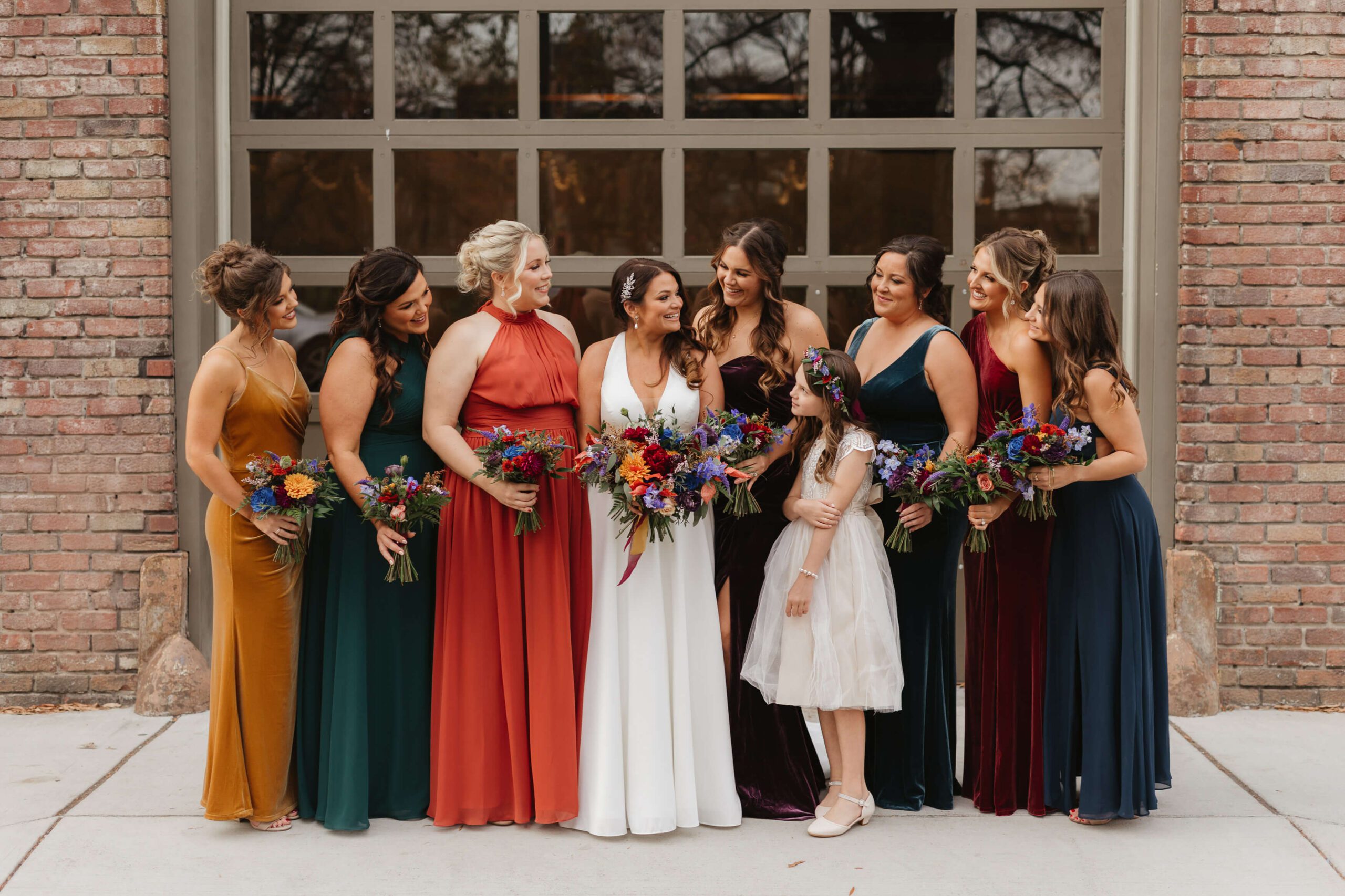 A bride in a white dress stands with her bridesmaids, who are wearing dresses in various colors. They are outside in front of a brick building, smiling and holding vibrant bouquets. A young girl stands among them in a light dress.