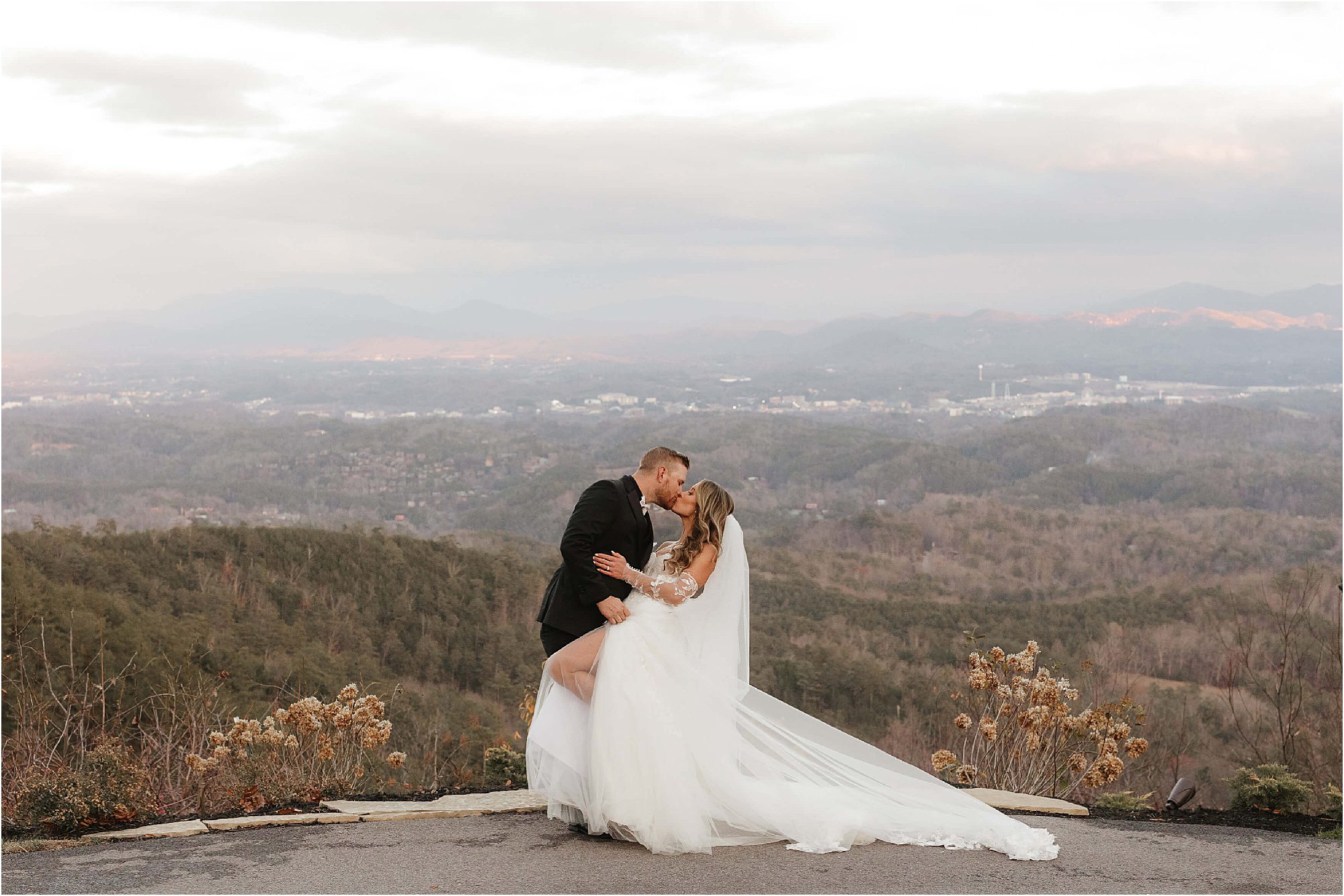 At The Trillium Venue, a bride and groom share a kiss on a scenic overlook. With mountains towering under a cloudy sky, the brides white dress contrasts elegantly with the grooms dark suit. Lush shrubs and foliage add charm to the picturesque setting.