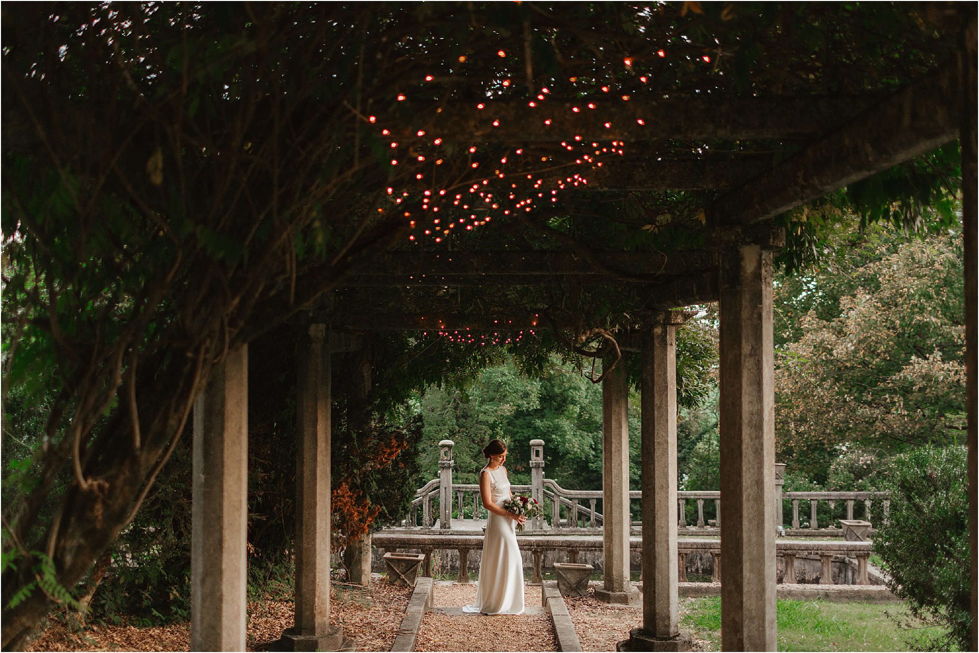 A person in a white dress stands under the vine-covered pergola at Bleak House Knoxville, adorned with twinkling lights. They clutch a bouquet, facing a stone pathway enveloped by lush greenery. A charming wooden bridge can be seen in the background, completing the enchanting scene.