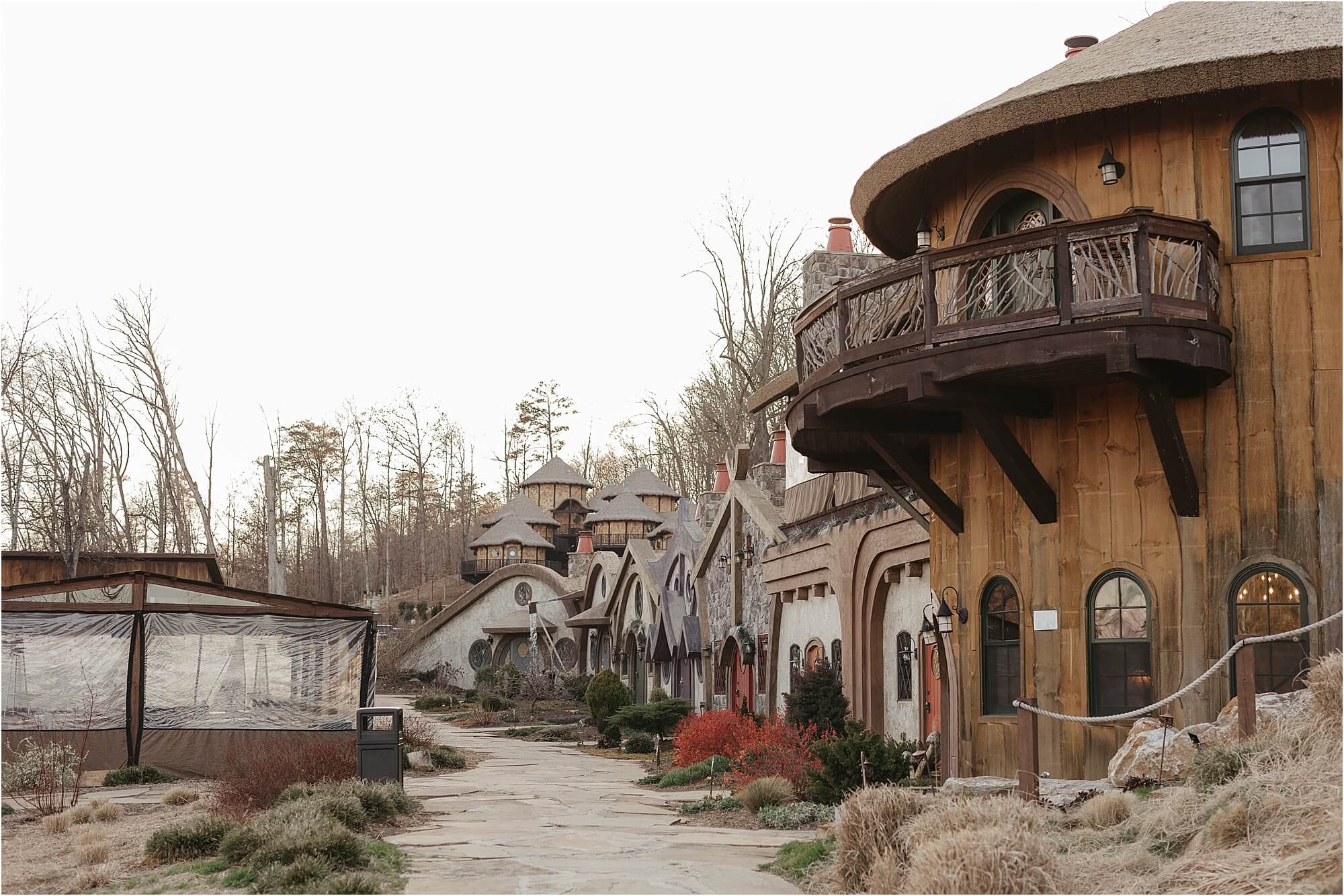 A village with round, hobbit-like houses featuring wooden and stone architecture. Theres a pathway lined with lanterns, surrounded by shrubs and grass. The scene gives a whimsical, fairytale vibe, with overcast skies and bare trees in the background.