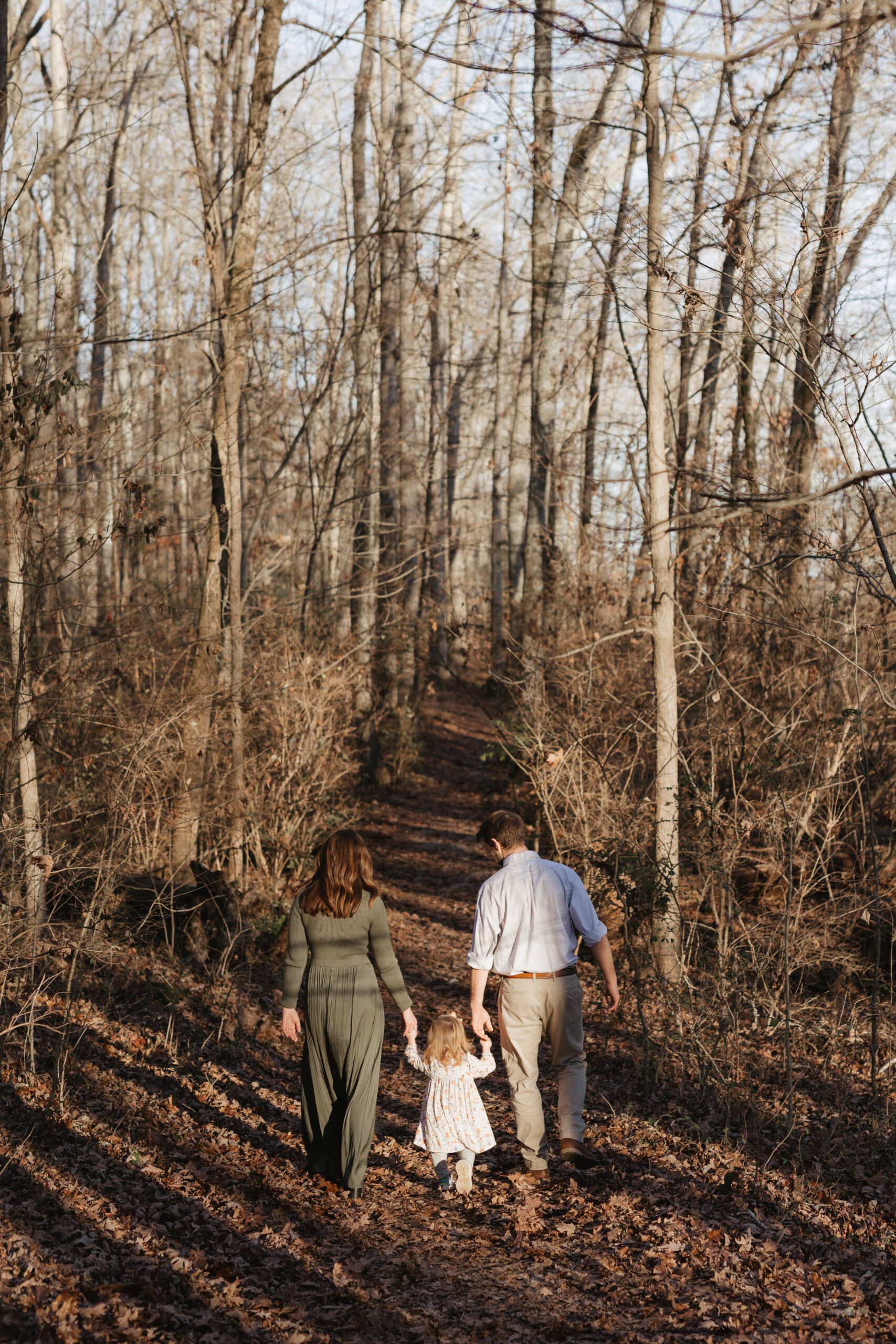 A family of three, including a small child, walks hand in hand along a leaf-covered forest trail. The trees are tall and leafless, indicating late autumn or early spring. Sunlight filters through the branches, casting long shadows on the ground.