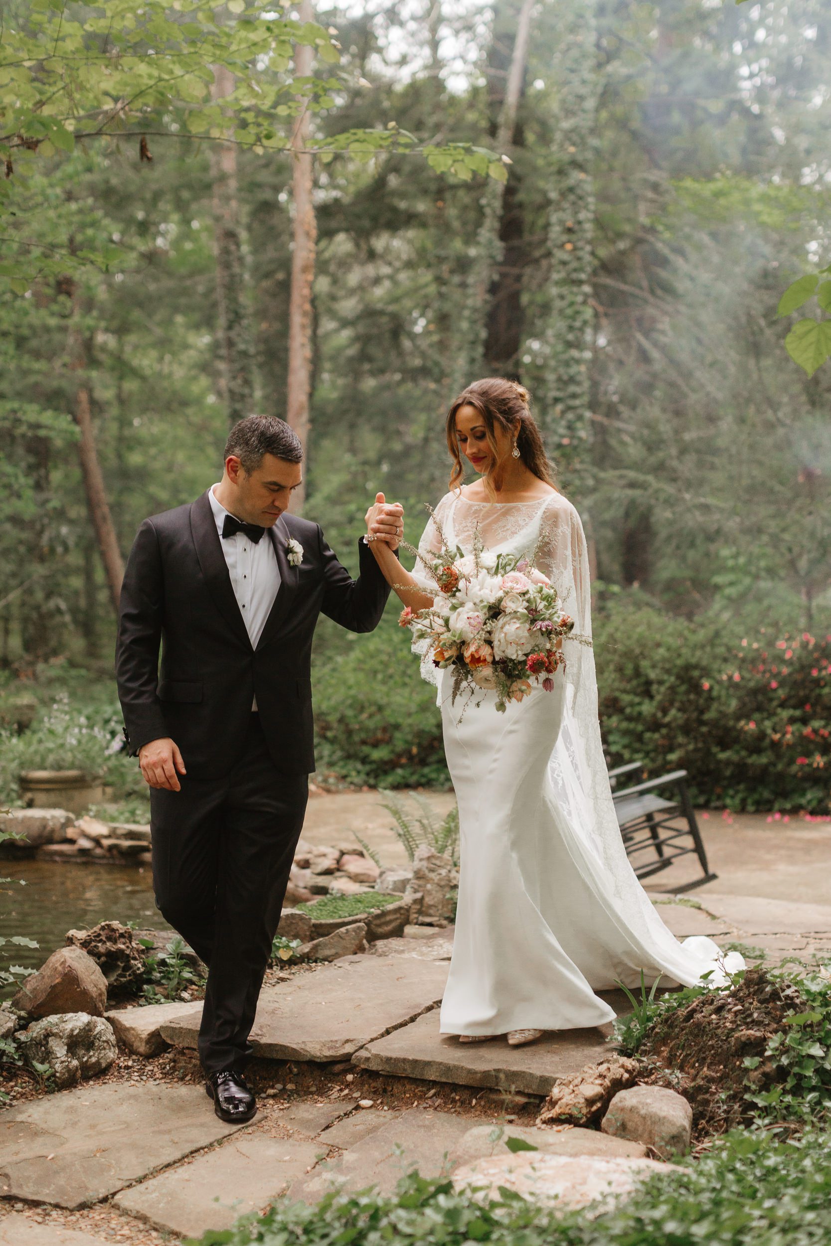 A groom in a black tuxedo holds hands with a bride in a white gown and veil as they walk down a stone path in a lush garden. The bride carries a bouquet of white and pink flowers. Trees and greenery surround them, with a small pond visible nearby.