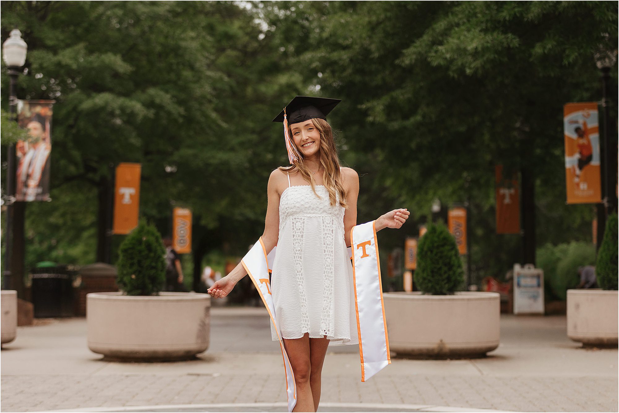 A young woman in a white dress and graduation cap smiles, standing outdoors on a campus walkway with orange banners and greenery in the background. She holds a white graduation stole with orange trim and a T on each end.