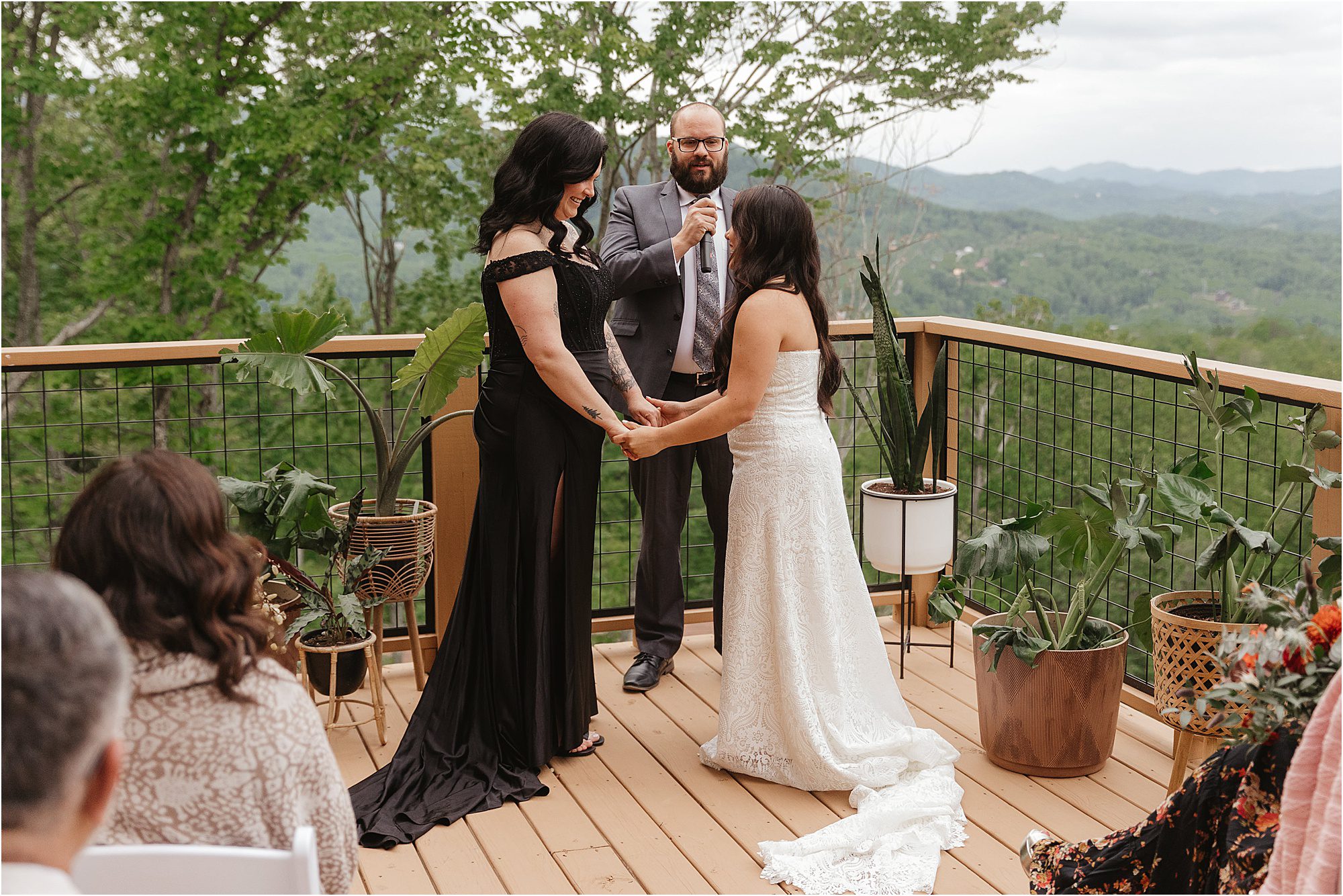 Two women stand holding hands during a wedding ceremony on the wooden deck at The Lunosa, surrounded by potted plants. With trees and mountains in the background, guests are seated as a man officiates the heartfelt moment.