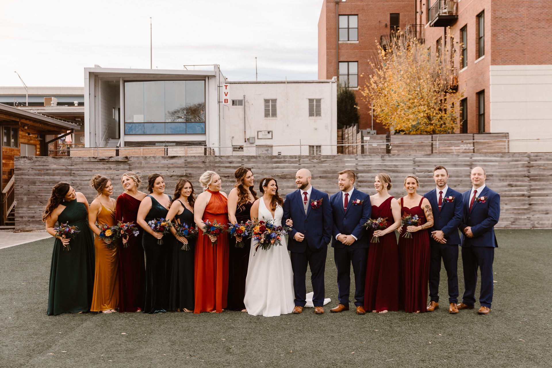 A wedding party poses outdoors at the Mill and Mine in formal attire. The bride stands center in white, surrounded by bridesmaids in colorful dresses and groomsmen in navy suits with red ties. Urban buildings and autumn trees frame the scene.