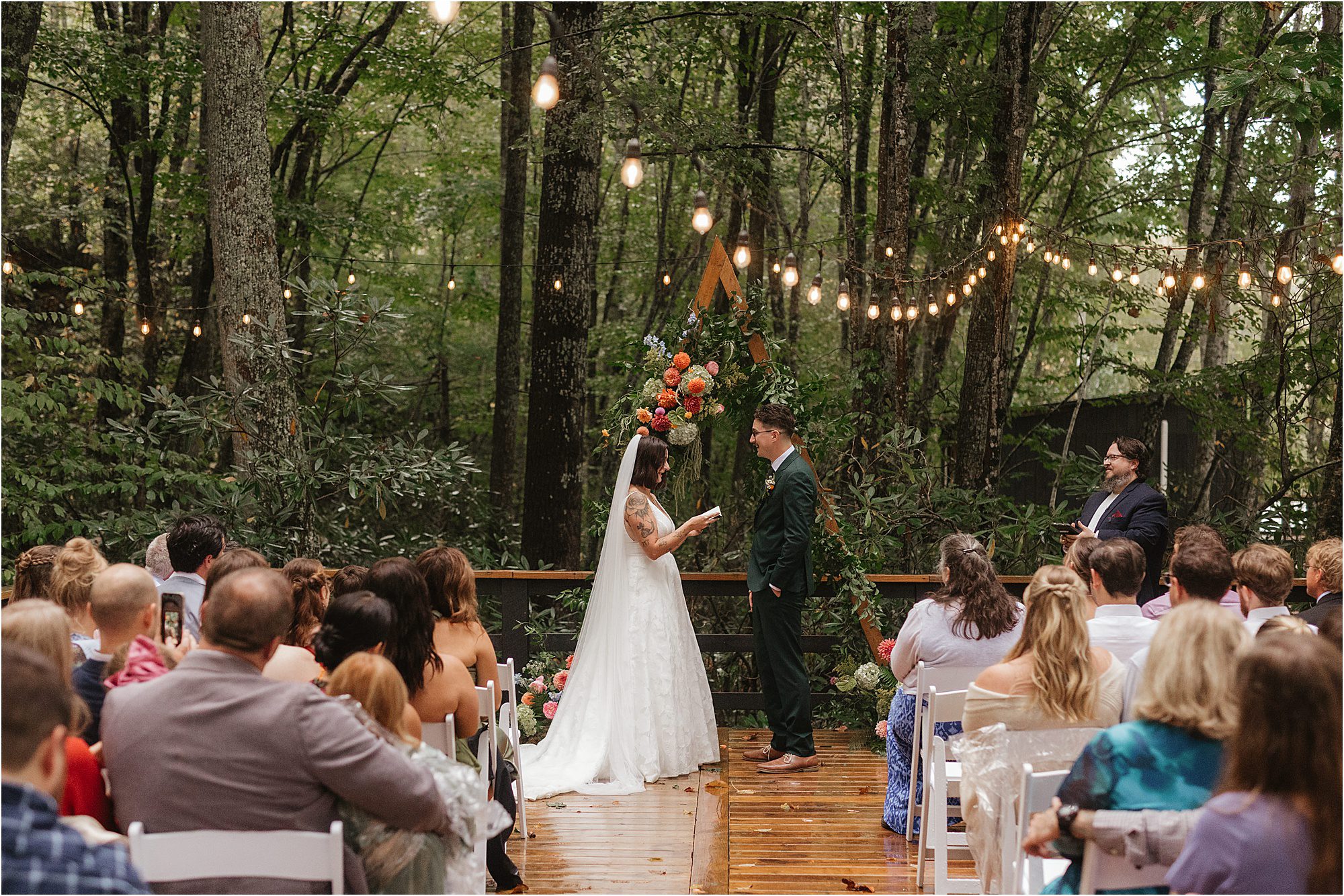 A bride and groom stand facing each other on a wooden platform in a forest, exchanging vows under retro string lights, with guests seated and a floral arrangement behind them, capturing the charm of a glamping-inspired celebration.