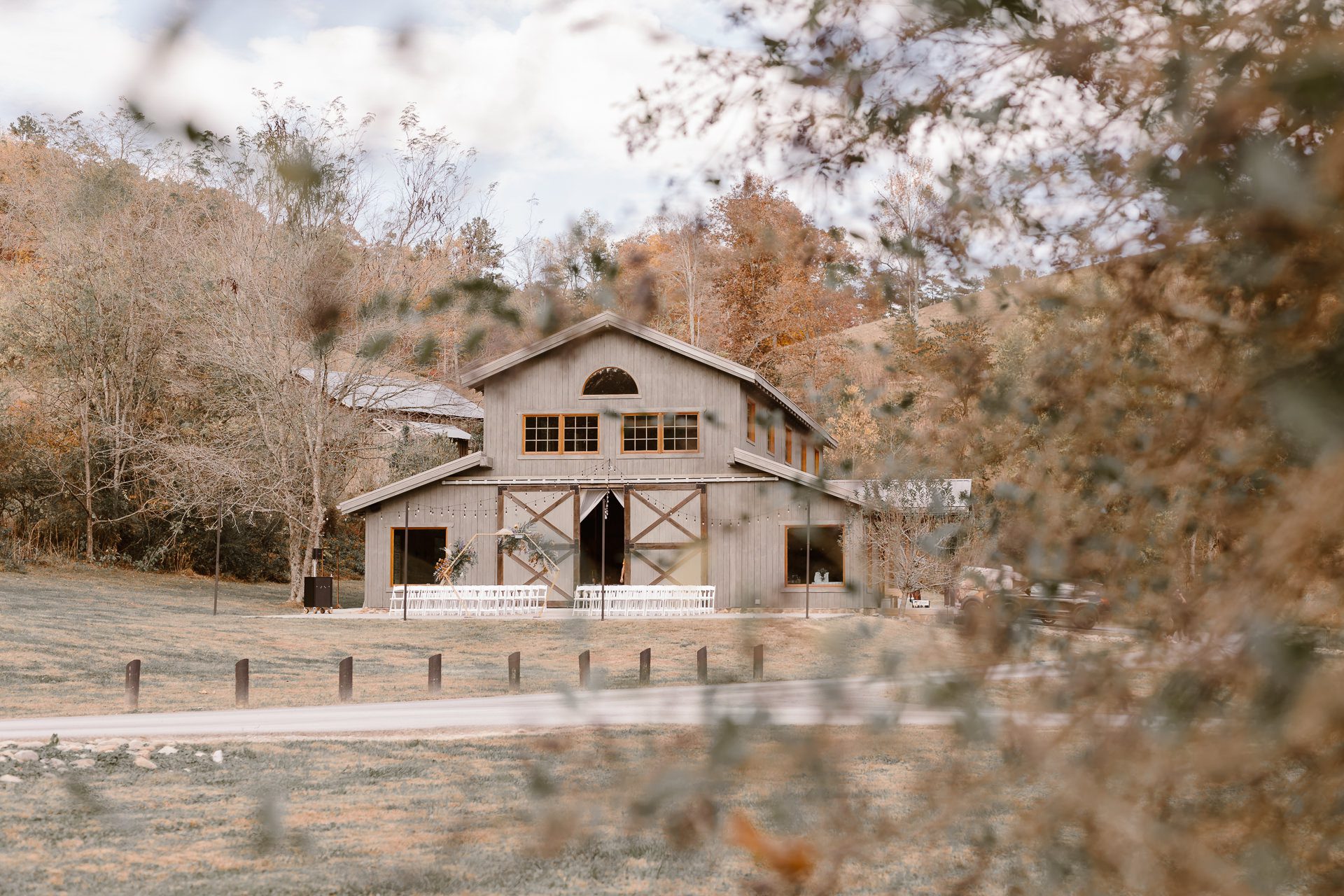 A rustic wooden barn with large windows sits among autumn trees and grass, partially obscured by blurred branches in the foreground. The scene has a warm, peaceful, countryside ambiance.