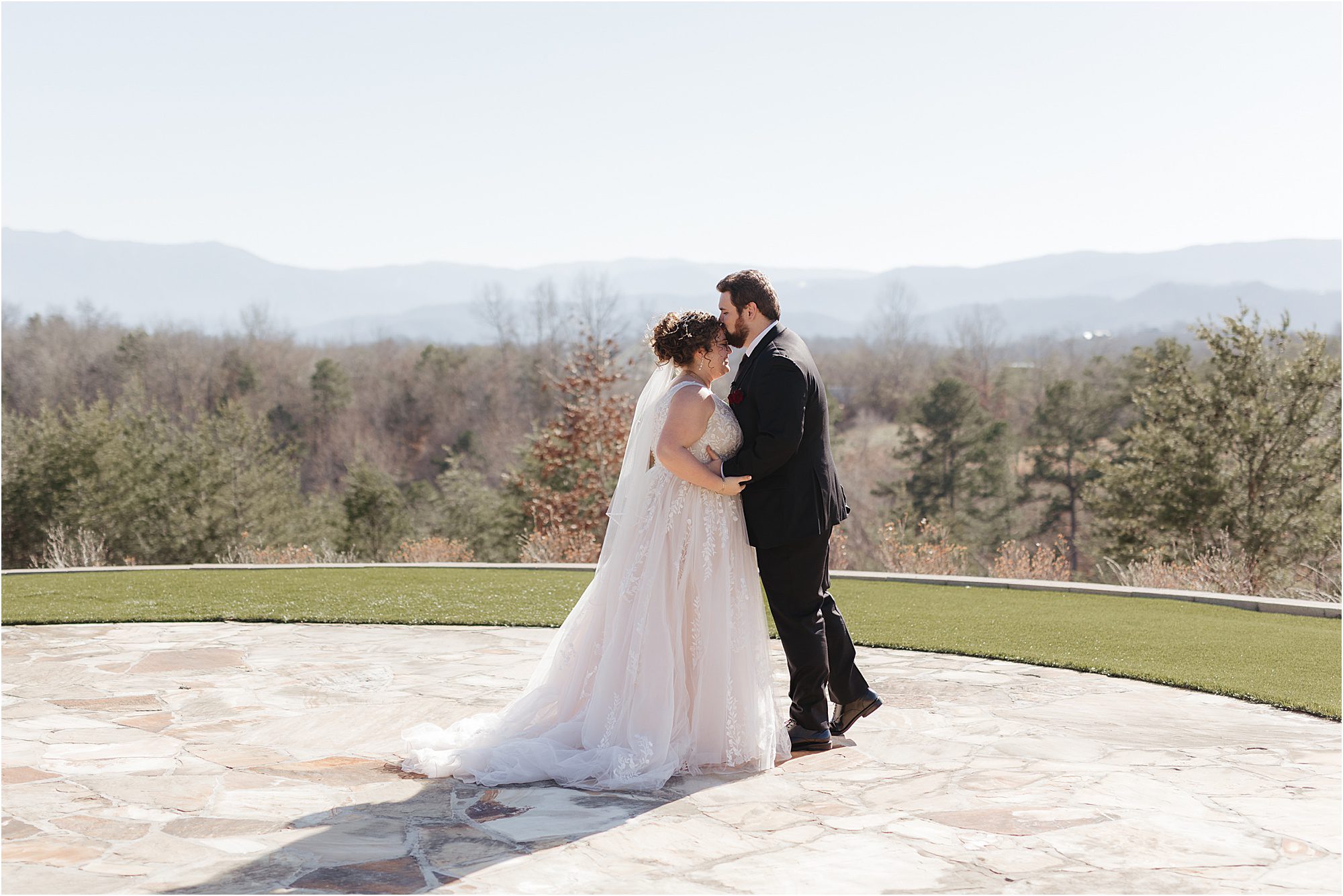 A bride and groom stand close together outdoors on a stone patio at Nichols Heir Wedding Venues, with trees and distant mountains in the background under a clear sky.