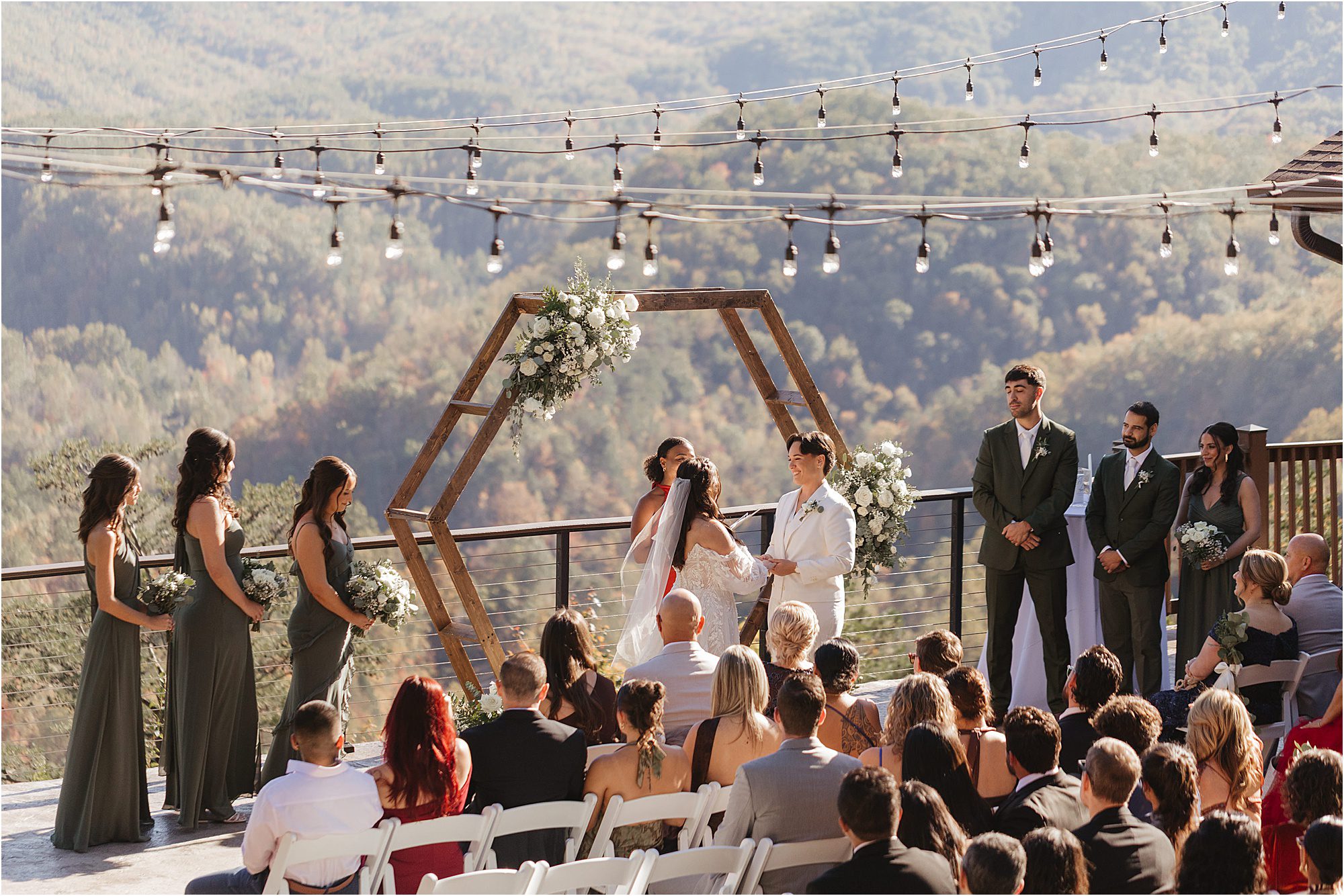 A wedding ceremony outdoors with mountain views; a couple stands under a hexagonal floral arch in green and cream wedding colors, exchanging vows, surrounded by their wedding party and seated guests. String lights are hung above.