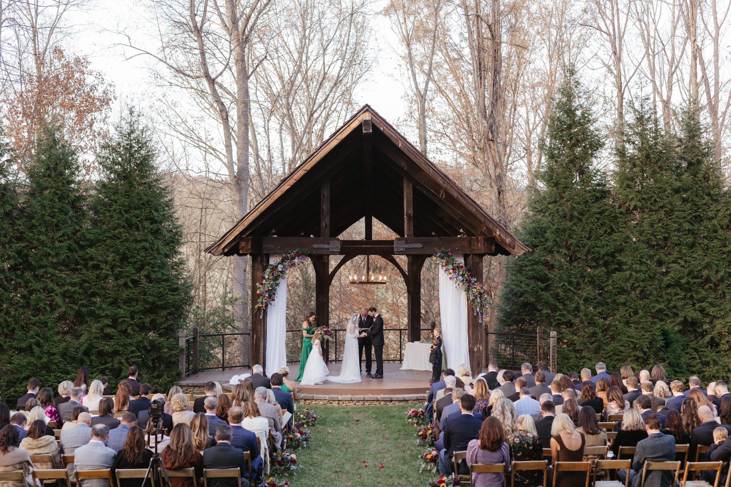 An outdoor wedding ceremony with a bride and groom standing under a wooden pavilion, surrounded by guests seated on either side, with tall trees and greenery in the background.