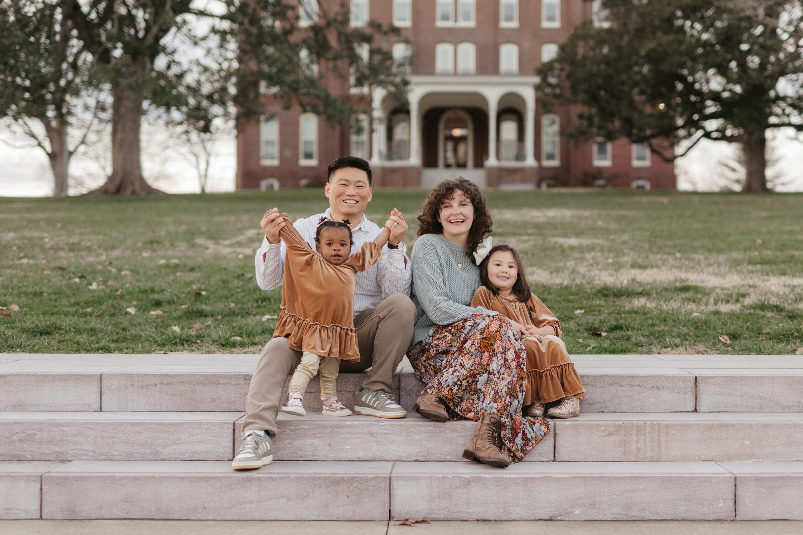 A smiling family of four sits on stone steps outdoors, with a large brick building behind them. The parents each hold a child, and everyone is dressed in warm, earth-toned clothing. Trees surround the lawn in the background.