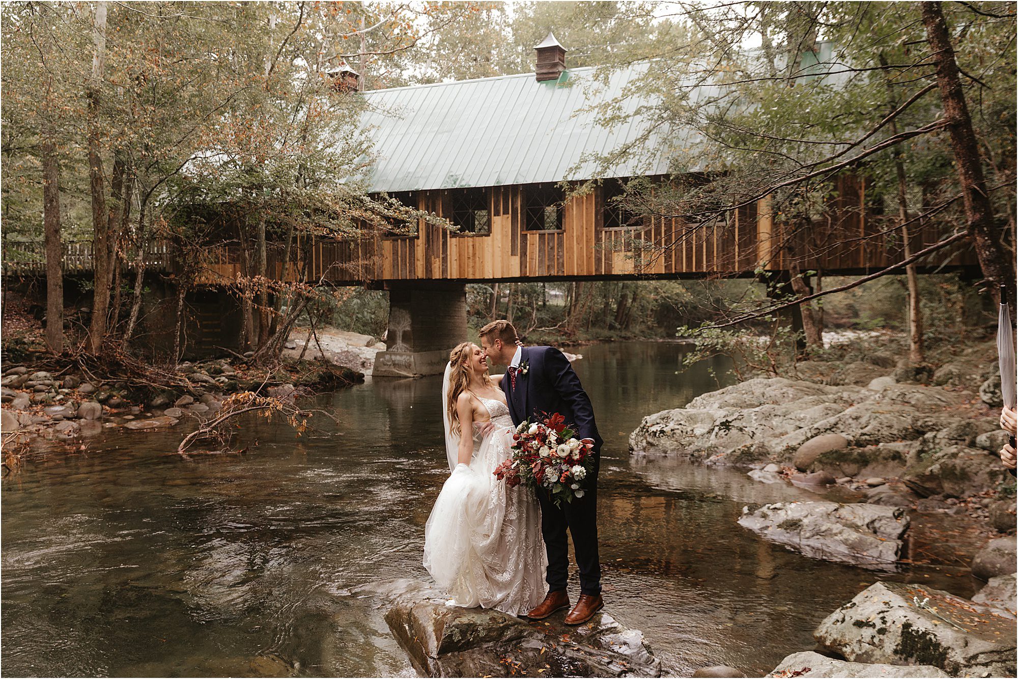 A bride and groom stand on rocks in a shallow creek at Greenbrier in the Great Smoky Mountains National Park, sharing a kiss. The bride holds a bouquet, and a rustic wooden covered bridge spans the water behind them, surrounded by trees.