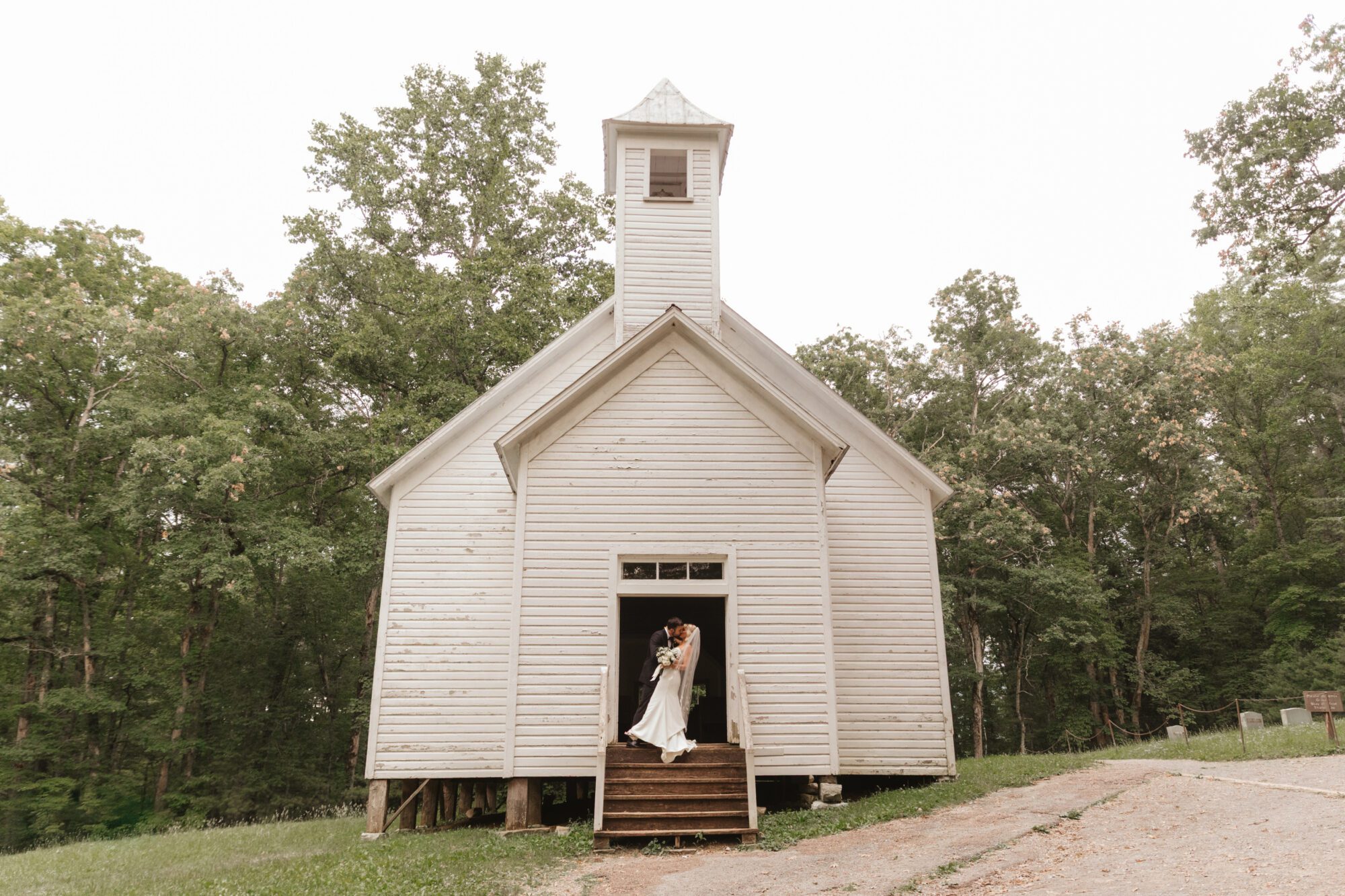 A bride and groom kiss in the doorway of a small, rustic white church surrounded by green trees under an overcast sky—one of the charming Cades Cove Churches.