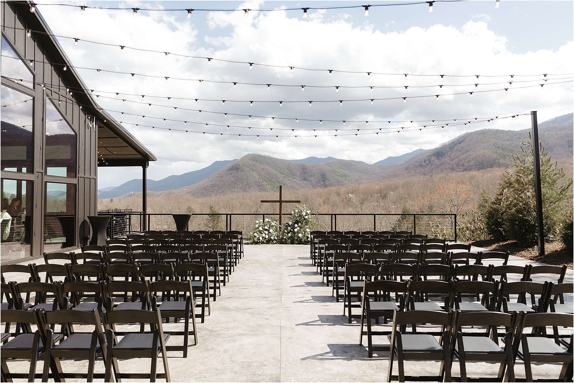 Rows of black chairs are arranged facing a floral cross on an outdoor patio at Trailhead Venues, with string lights overhead and scenic mountains in the background under a partly cloudy sky.