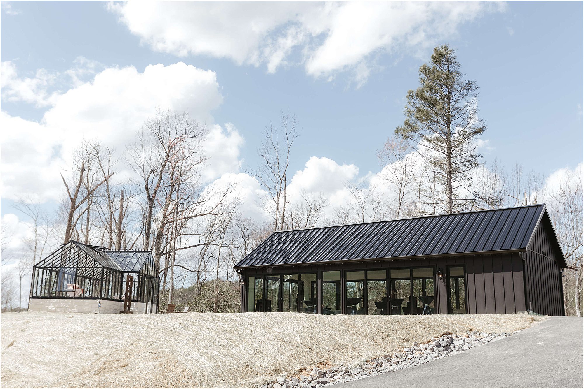 A modern black building with large windows sits beside a glass greenhouse on a grassy hill at Trailhead Venues, surrounded by bare trees under a partly cloudy sky.