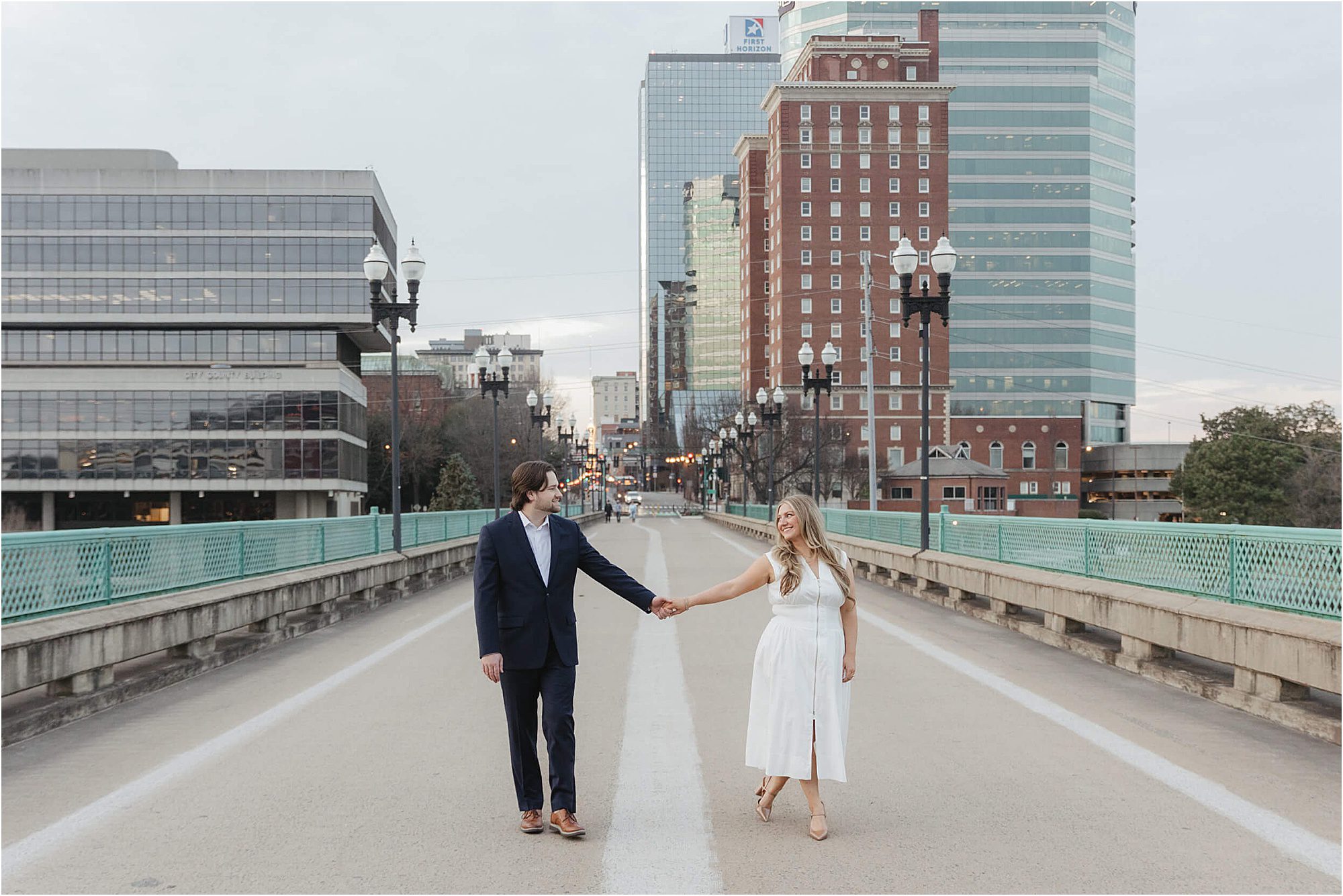 A man and woman holding hands stand in the center of an empty city street, dressed in semi-formal attire, reminiscent of Gay Street Bridge engagement photos, with tall buildings and streetlights in the background.