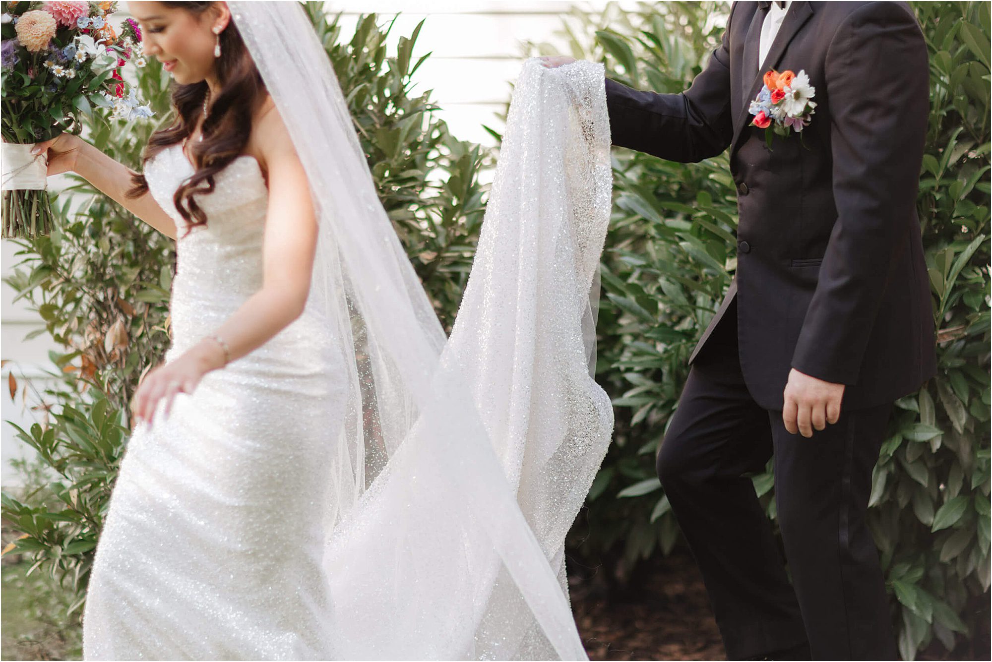 A bride in a sparkly white gown holds a bouquet while a groom in a black suit carries her long veil. They are walking outdoors, surrounded by greenery, capturing the natural beauty of a wedding in East Tennessee.