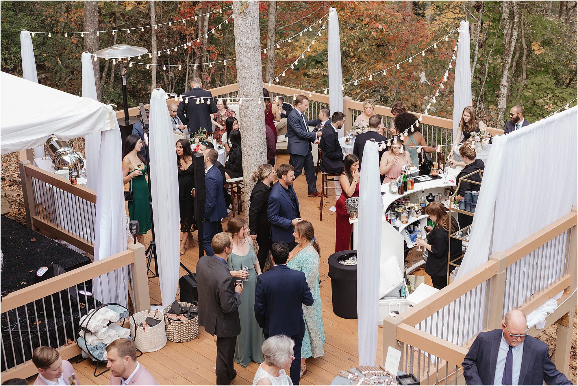 Guests in formal attire mingle on a wooden deck at an outdoor event, exemplifying wedding ideas that keep guests engaged. String lights hang overhead as people gather near the bar, surrounded by trees and vibrant autumn foliage in the background.