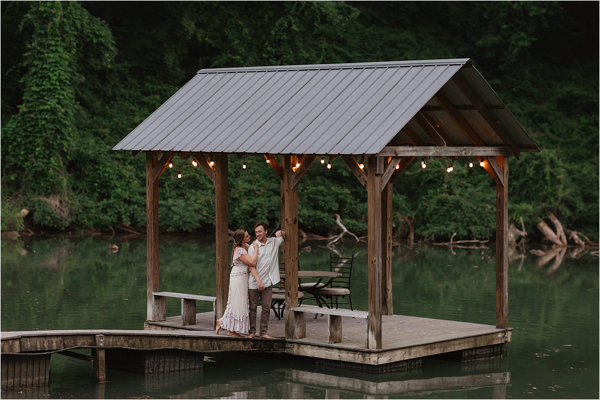 A couple stands close together under a wooden dock shelter with string lights, surrounded by calm water and lush green trees in the background, creating a dreamy lakeside engagement session.