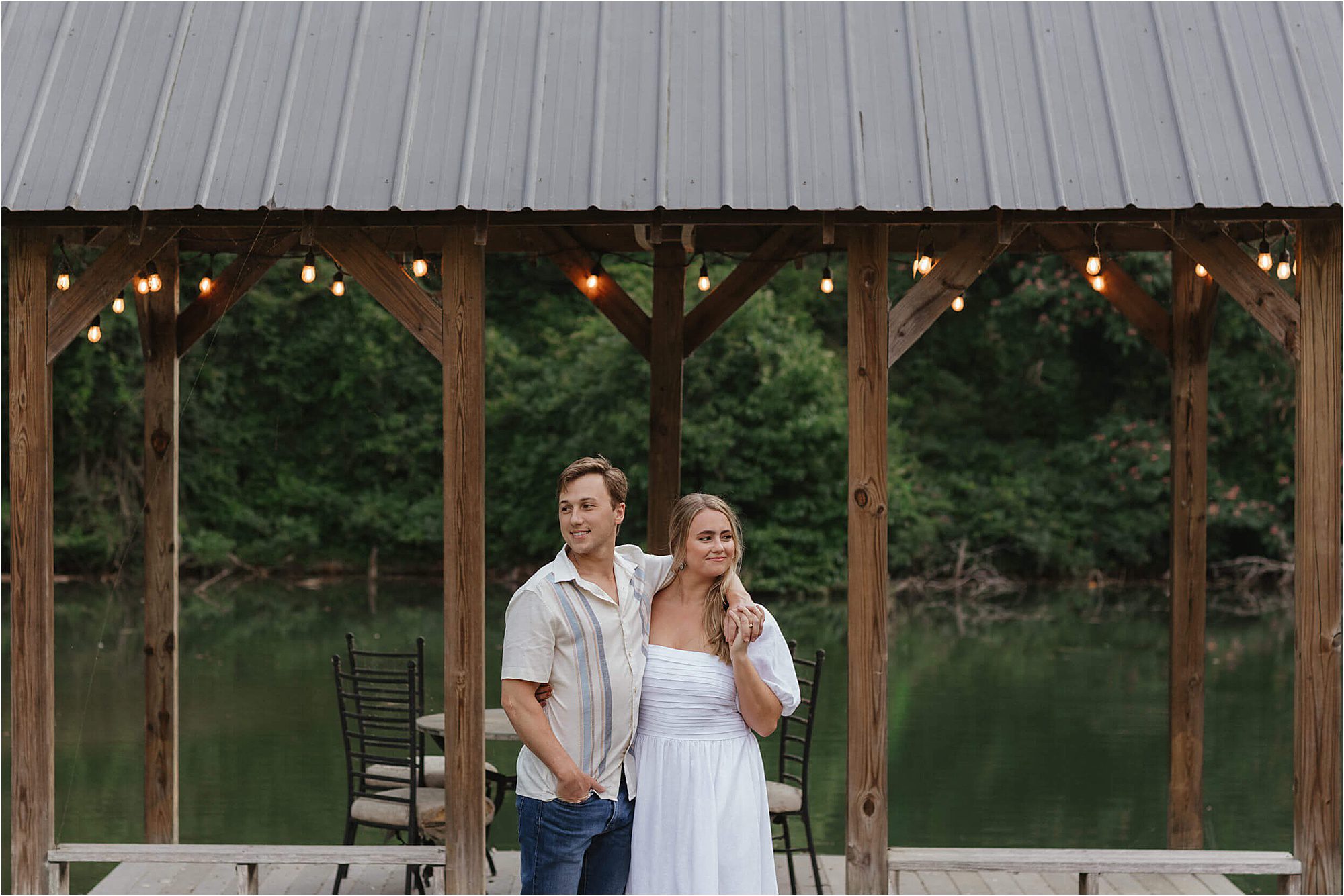 A couple stands under a wooden pavilion with string lights by a lake, surrounded by trees during their lakeside engagement session. The woman wears a white dress and the man wears a striped shirt and jeans.