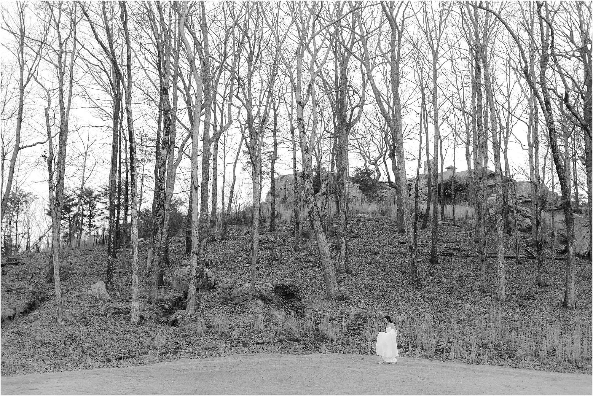 A person in white sits on the ground at the edge of a leafless forest, with tall, bare trees and a rocky hill in the background. The peaceful scene, captured in black and white, evokes the quiet reflection that might surround a wedding at Cloudland at McLemore Resort.