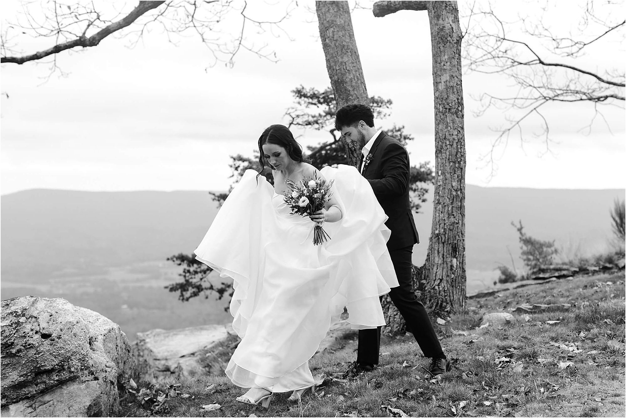 A bride holding flowers walks in a grassy, rocky area with a groom following behind her during their wedding at Cloudland at McLemore Resort. The scene is outdoors near trees and mountains, captured in black and white.