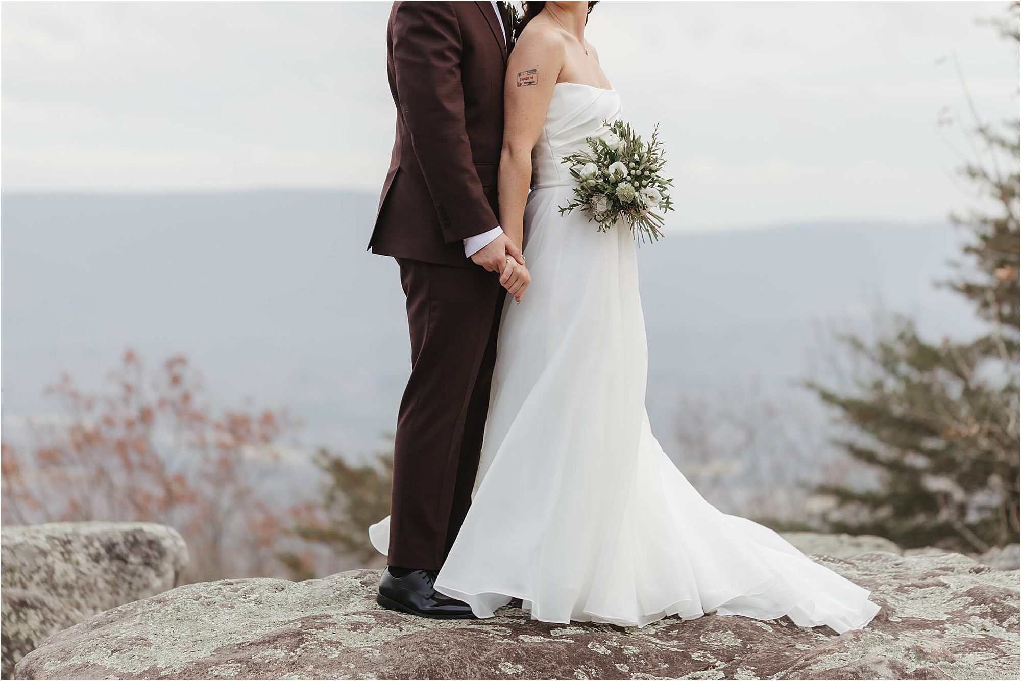 A bride in a white dress and a groom in a dark suit stand on a rock, holding hands during their wedding at Cloudland at McLemore Resort. The bride holds a bouquet with greenery. Their heads are not visible; mountains and trees are in the background.