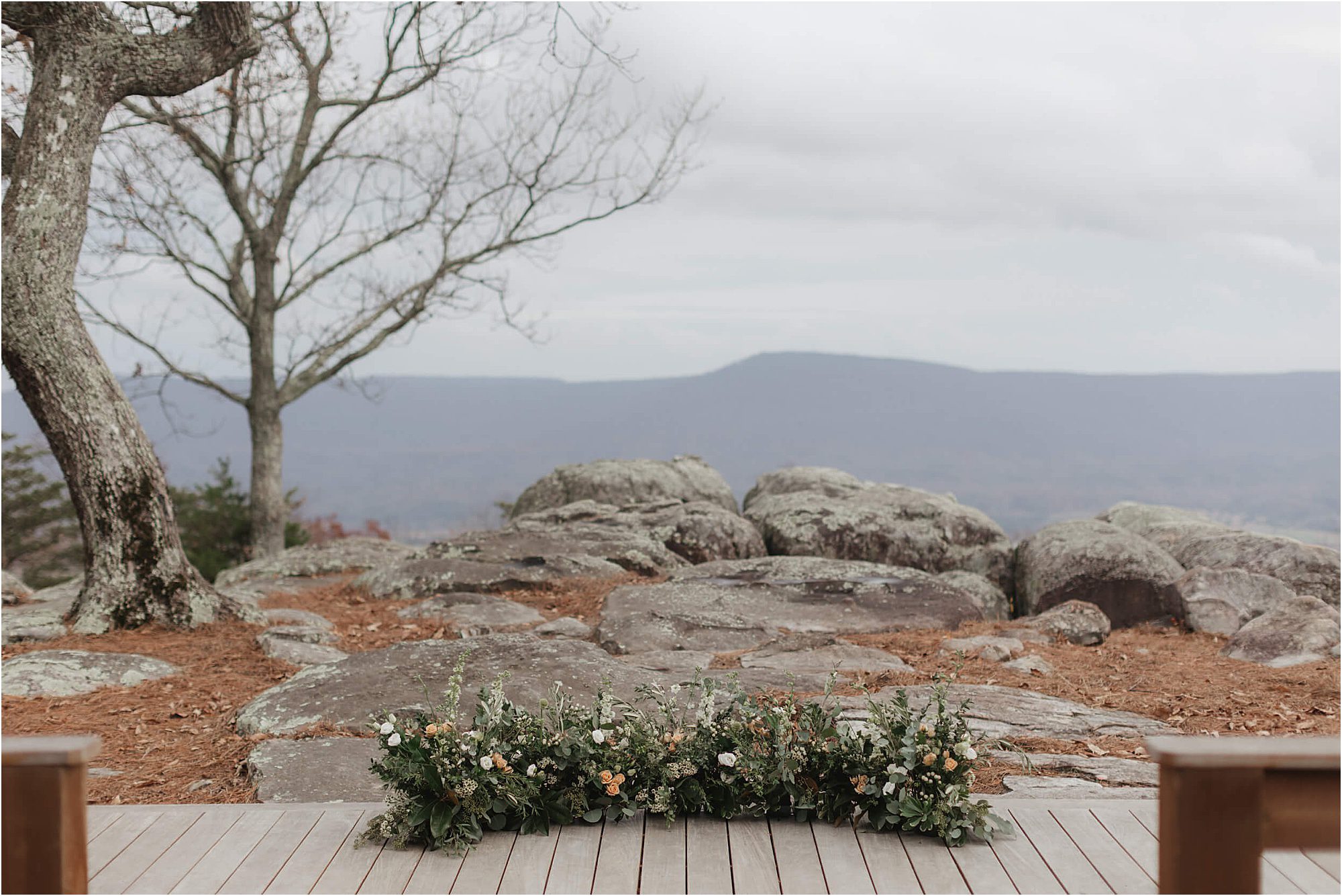 A wooden deck set for a wedding at Cloudland at McLemore Resort, adorned with a low floral arrangement, overlooks large gray rocks, bare trees, and distant mountains under a cloudy sky.