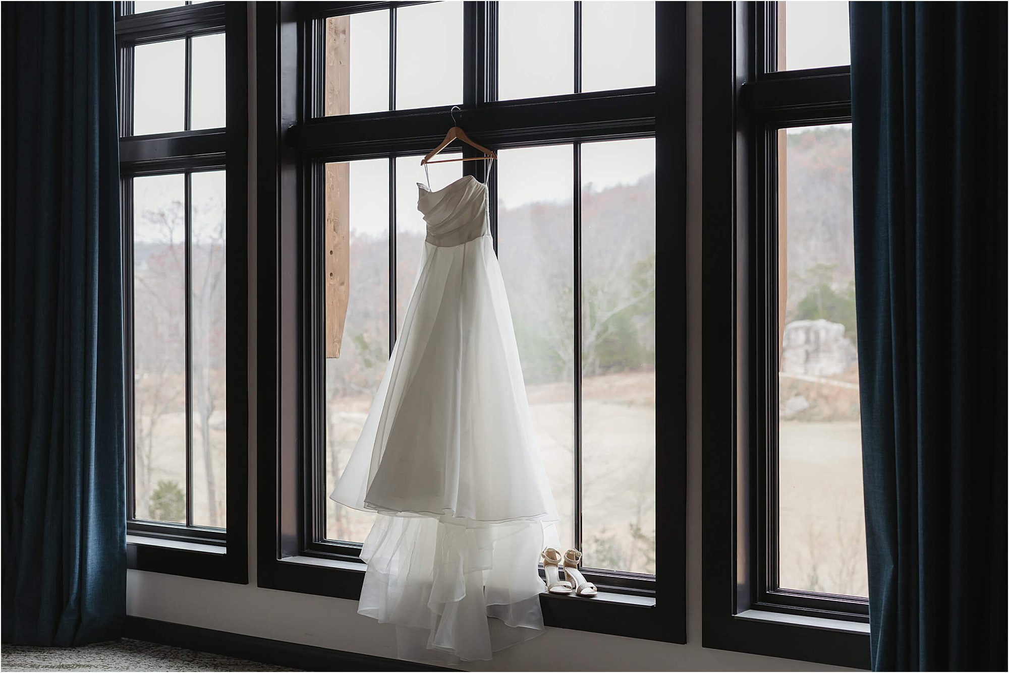 A white wedding dress hangs on a wooden hanger in front of large windows with blue curtains at a dreamy wedding at Cloudland at McLemore Resort; white shoes are placed neatly on the windowsill below.