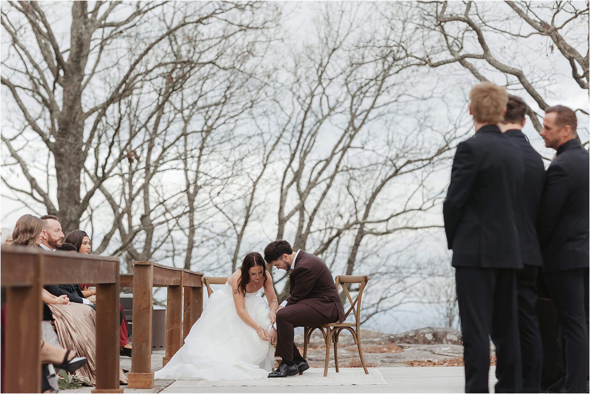 A bride and groom sit together outdoors during their wedding at Cloudland at McLemore Resort, heads bowed in an emotional moment, surrounded by their wedding party and guests, with leafless trees in the background.