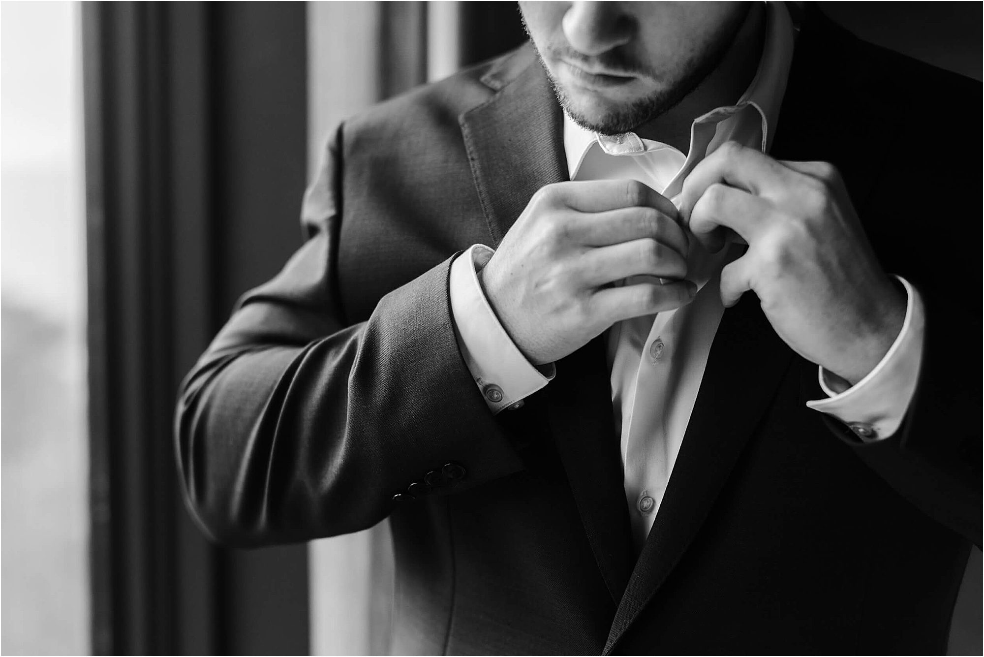 Black and white photo of a man in a suit buttoning his shirt collar near a window; his face is partially visible and he appears focused on getting dressed for a wedding at Cloudland at McLemore Resort.