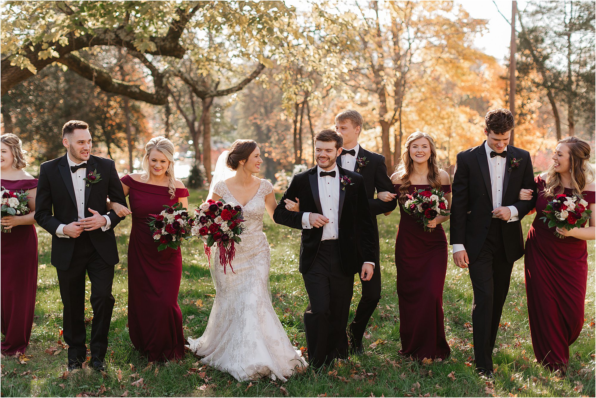 A bride and groom walk outdoors with their bridal party at Historic Westwood. The bridesmaids wear burgundy dresses and hold bouquets, while the groomsmen don black tuxedos. Sunlight and autumn trees fill the background, adding to the timeless charm of this celebrated venue.