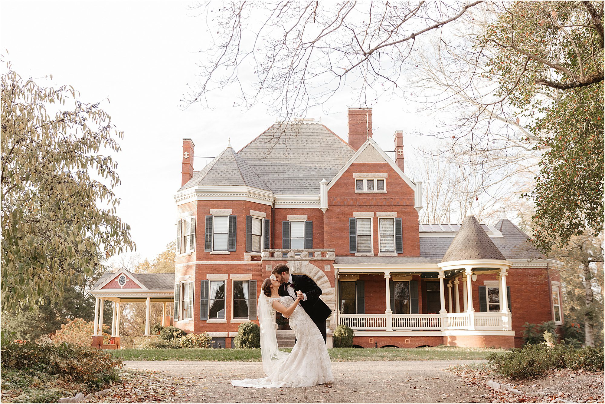 A groom dips and kisses a bride in a white gown in front of the elegant, historic Westwood mansion, with its wraparound porch and turret, surrounded by trees and beautiful landscaping.