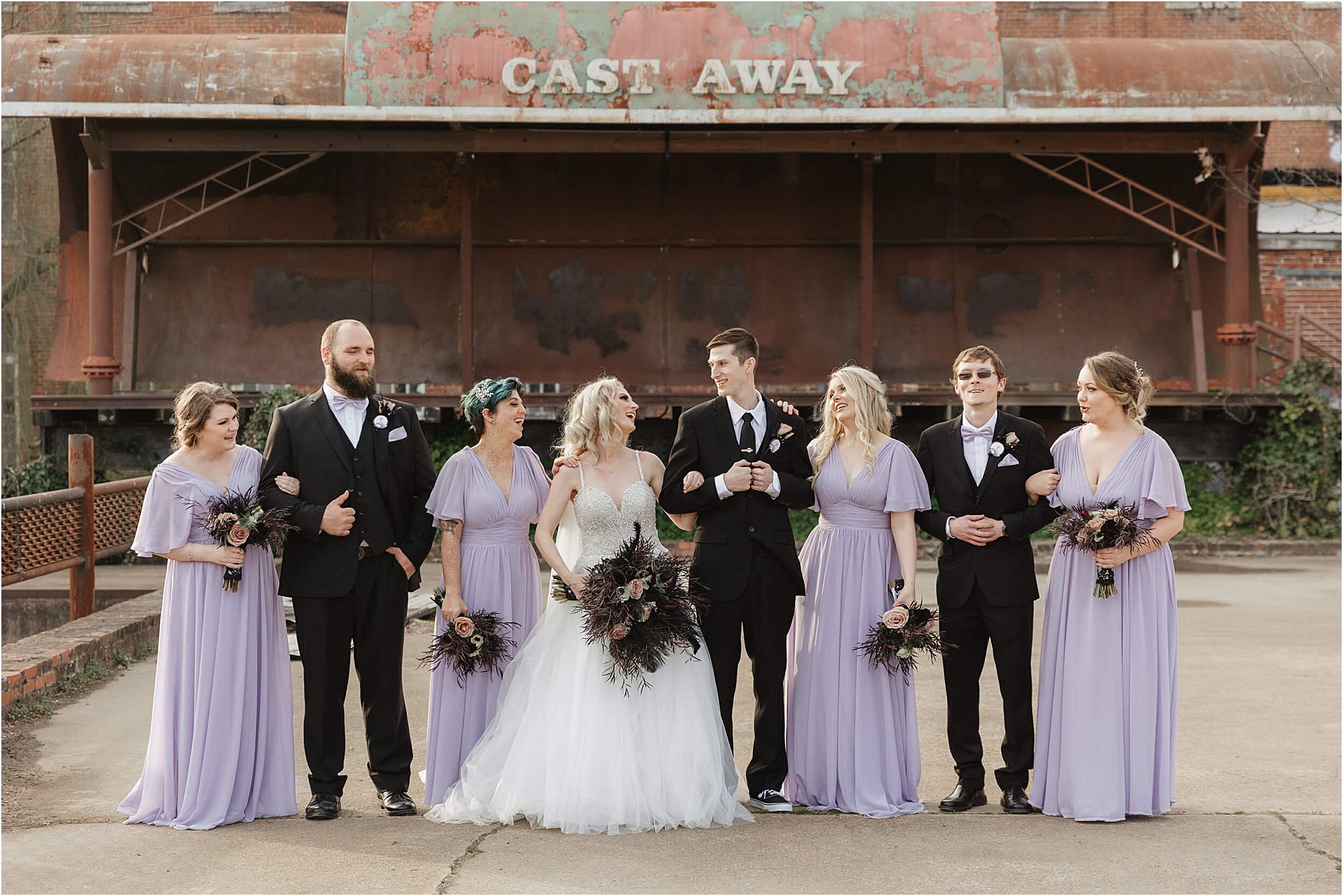 A wedding party poses outdoors in front of a weathered Cast Away sign near the Weaver’s Room. The bride wears a white gown, bridesmaids wear lavender dresses, and the groomsmen wear black suits. Everyone is smiling and linking arms.