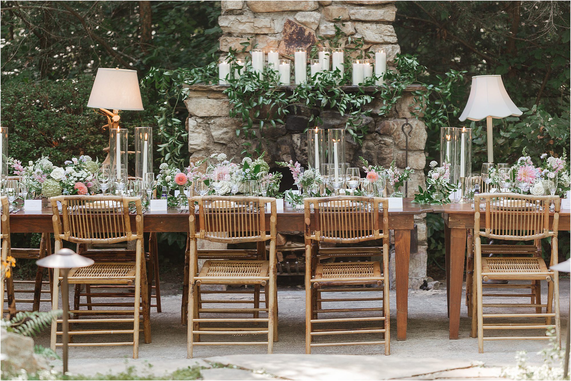 A long wooden table with bamboo chairs is set outdoors with floral centerpieces that showcase stunning wedding color combinations, complemented by glass candle holders and two lamps. Behind, a stone fireplace is decorated with greenery and large white candles.