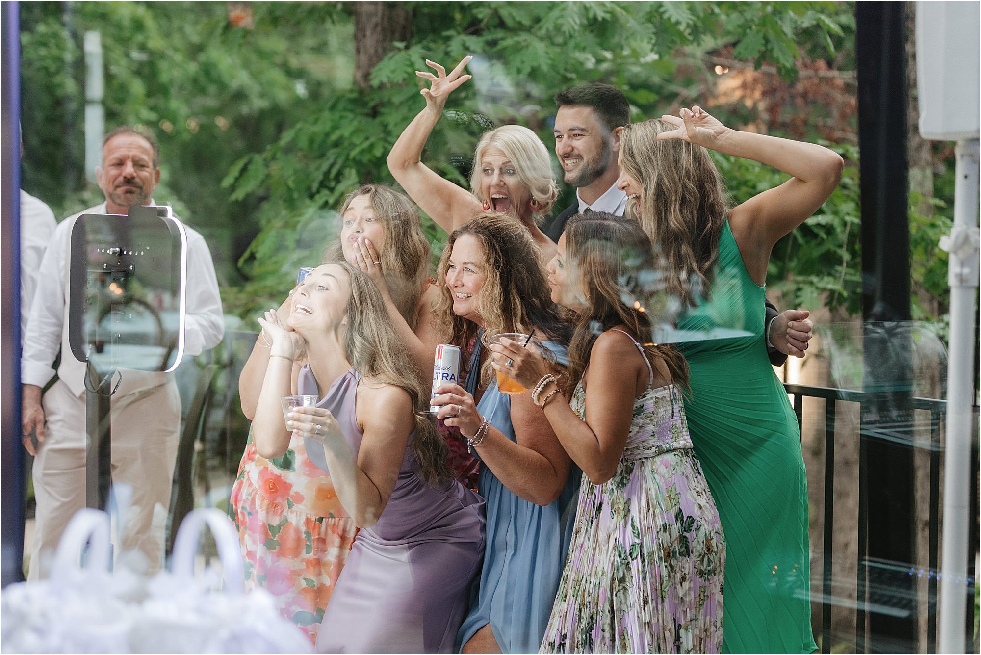 A group of people in colorful dresses and a suit pose playfully for a photo outdoors, holding drinks and making silly faces with greenery in the background—capturing one of those wedding ideas that keep guests engaged and having fun throughout the celebration.
