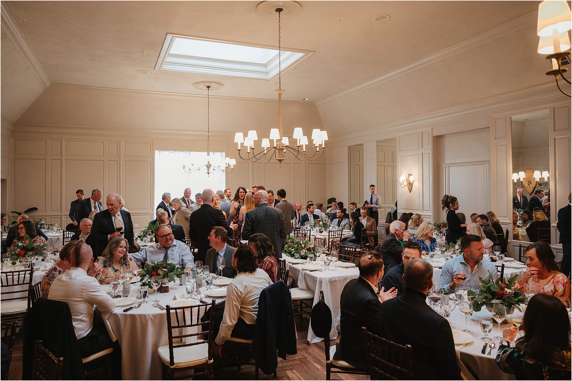 Guests sit and stand around round tables decorated with flowers in a bright, elegant banquet hall with chandeliers. People are talking, eating, and enjoying a formal event filled with wedding ideas that keep guests engaged throughout the celebration.