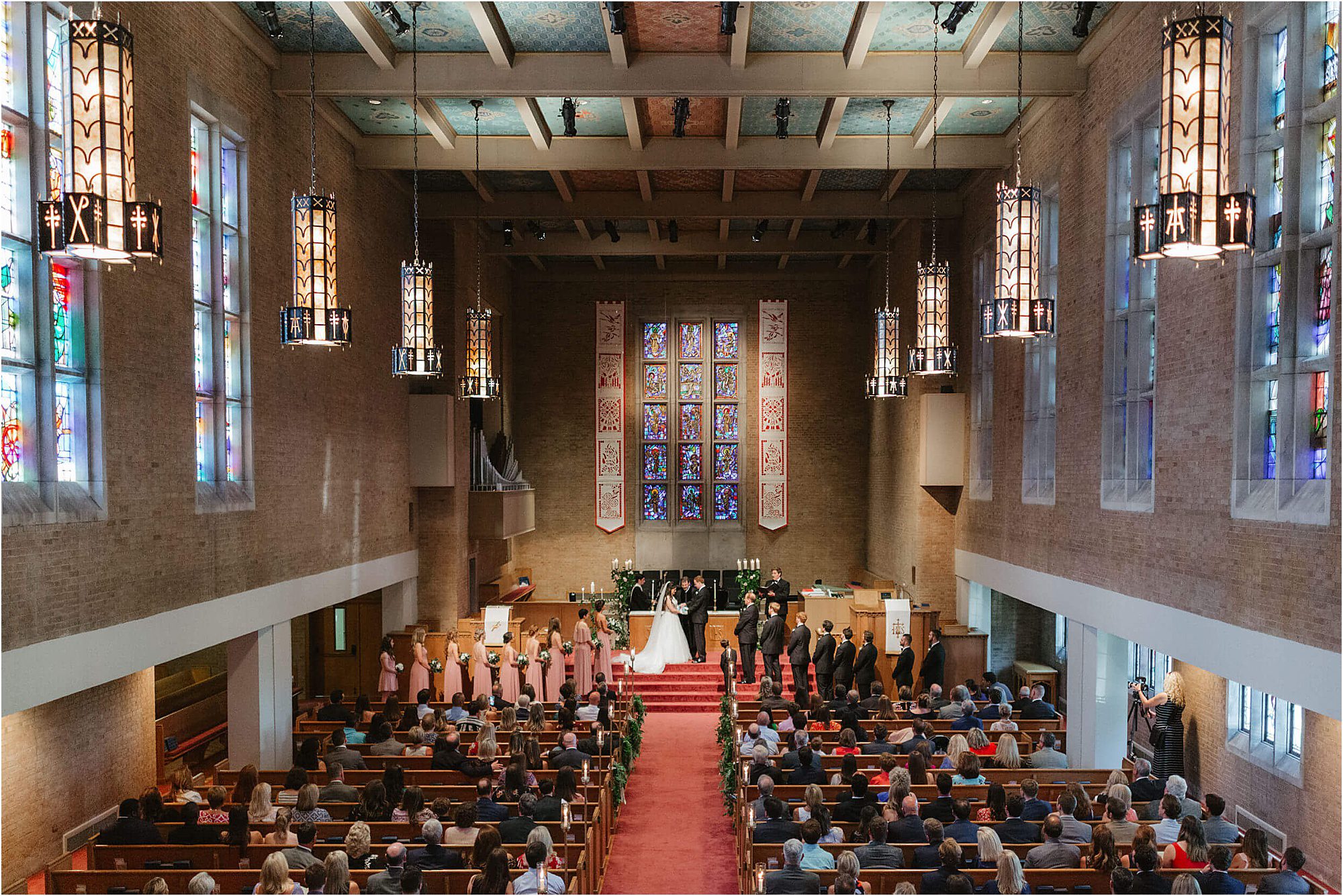 A wedding ceremony takes place at 2nd Presbyterian Church with stained glass windows, high ceilings, and rows of guests seated on wooden pews. The wedding party stands at the altar.