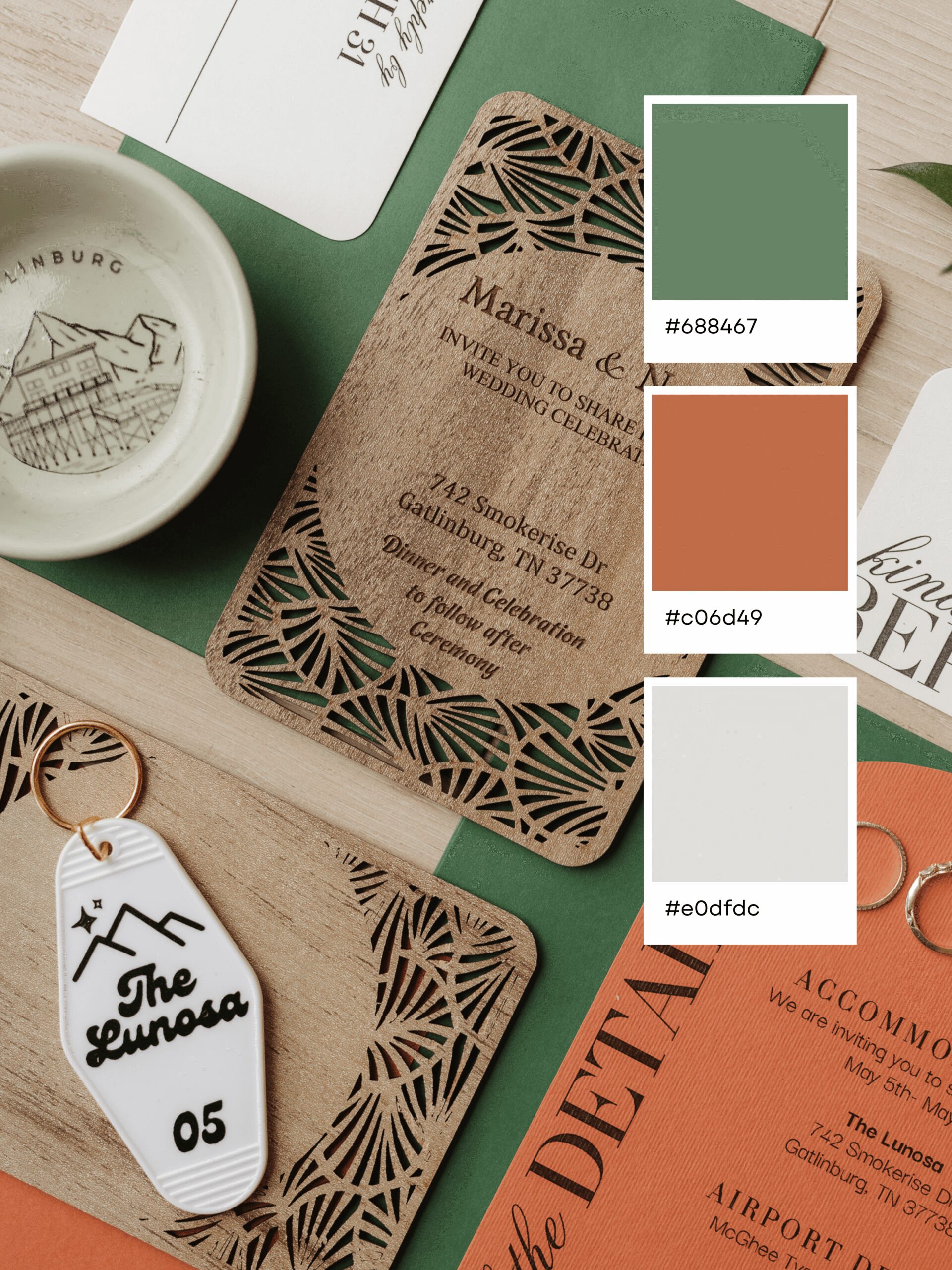 Flat lay of wedding invitations, a keychain with The Lunosa 05, a small dish, green envelope, and three color swatches—sage green (#688A67), orange (#C06D49), and off-white (#E0DFDC)—perfect for inspiring unique wedding color combinations.