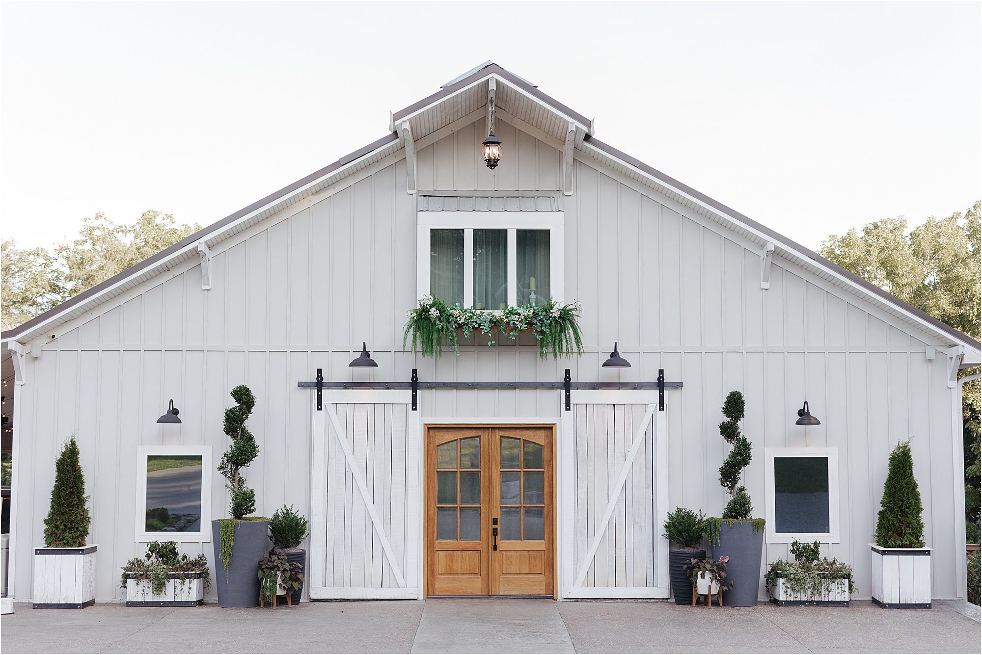 A modern white barn at Ramble Creek features wooden double doors, potted plants, and greenery above the entrance. Two sliding barn doors and three outdoor lights enhance the symmetrical design. Trees are visible in the background.